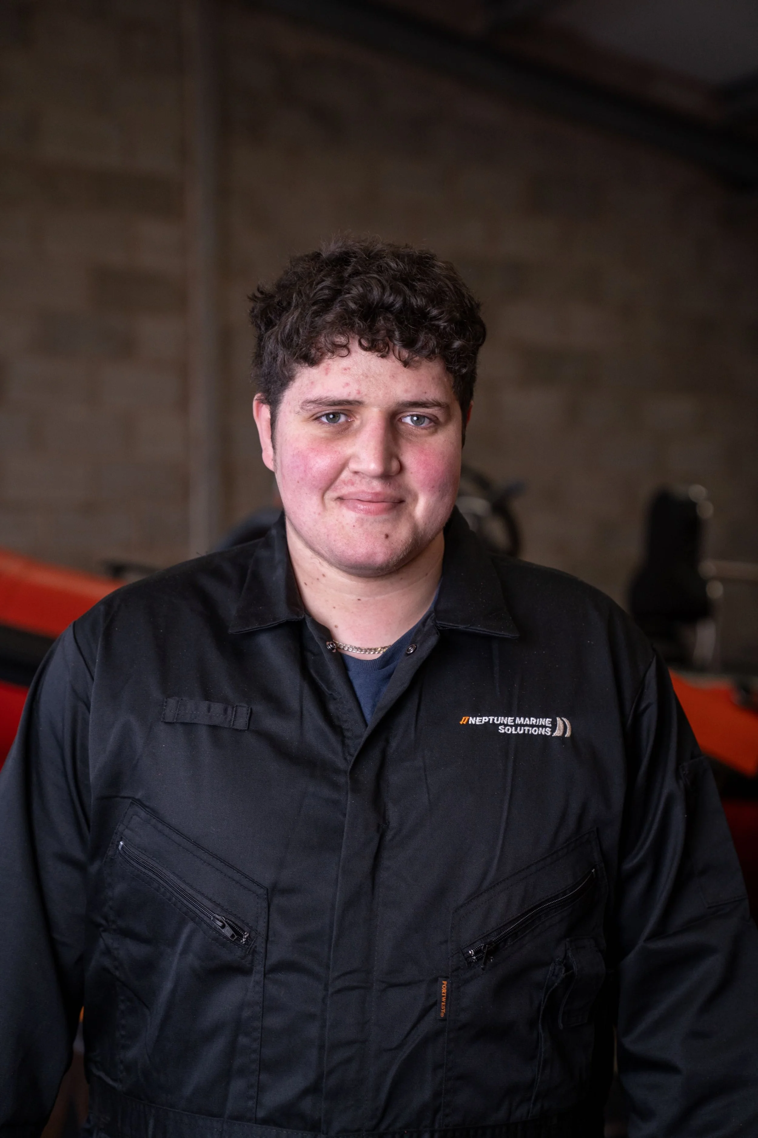 A young man with curly dark hair and a fair complexion posing indoors in front of a brick wall, wearing a black jacket with a logo that reads Neptune Marine Solutions.
