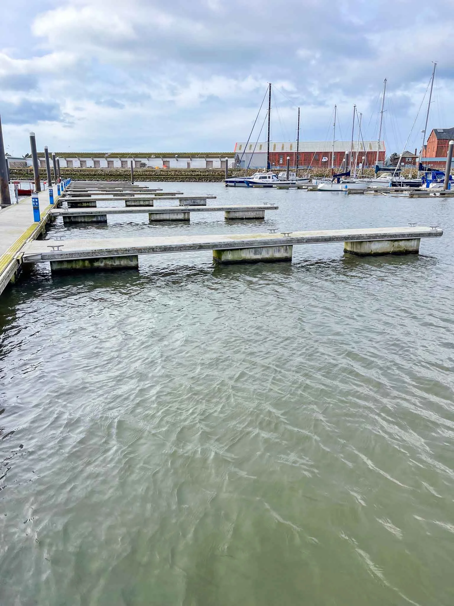 Marina with empty boat docks, sailboats moored in the background, and cloudy sky.