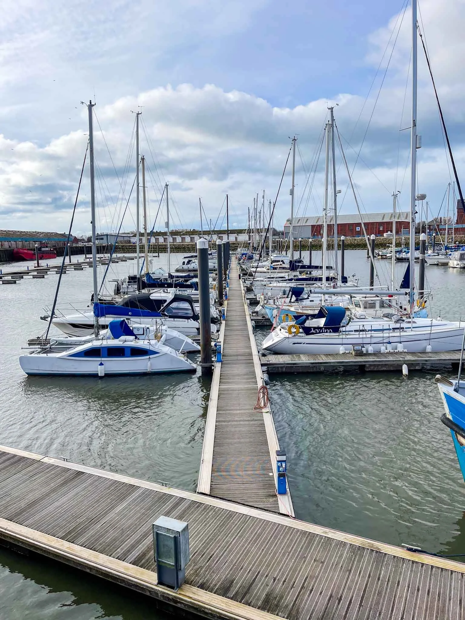 A marina with sailboats and yachts docked along wooden piers under a partly cloudy sky.