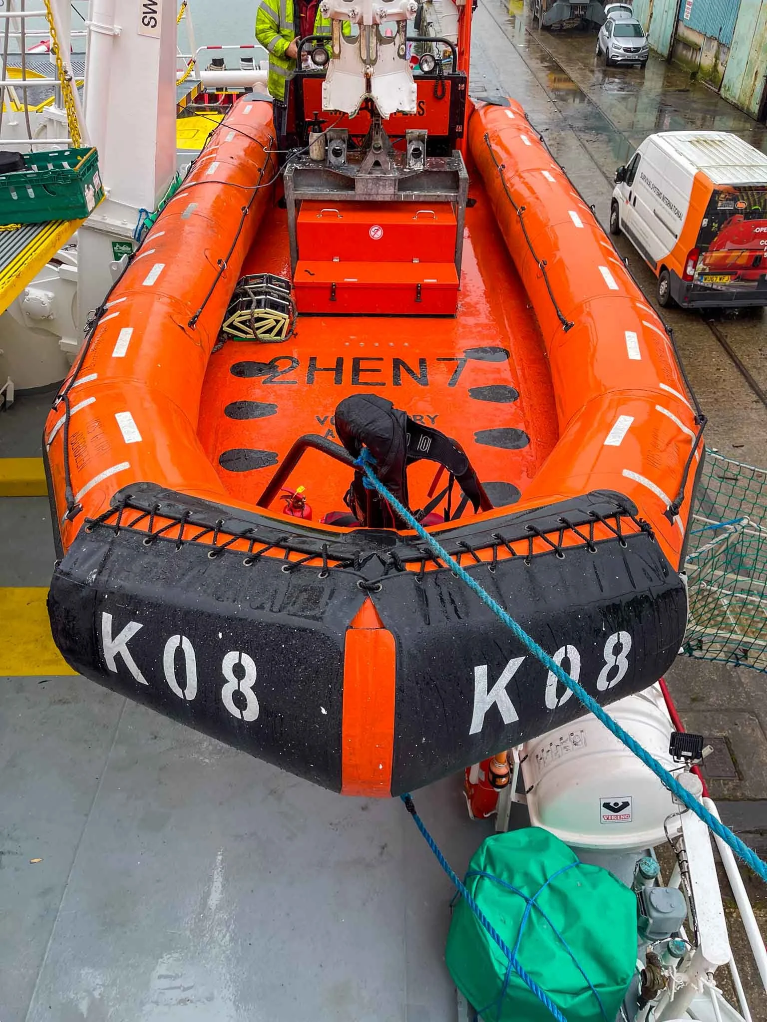An orange rescue boat with black and white markings, labeled 'K08', on a vessel deck during rainy weather. There are equipment and personnel aboard, with a street and vehicles visible in the background.