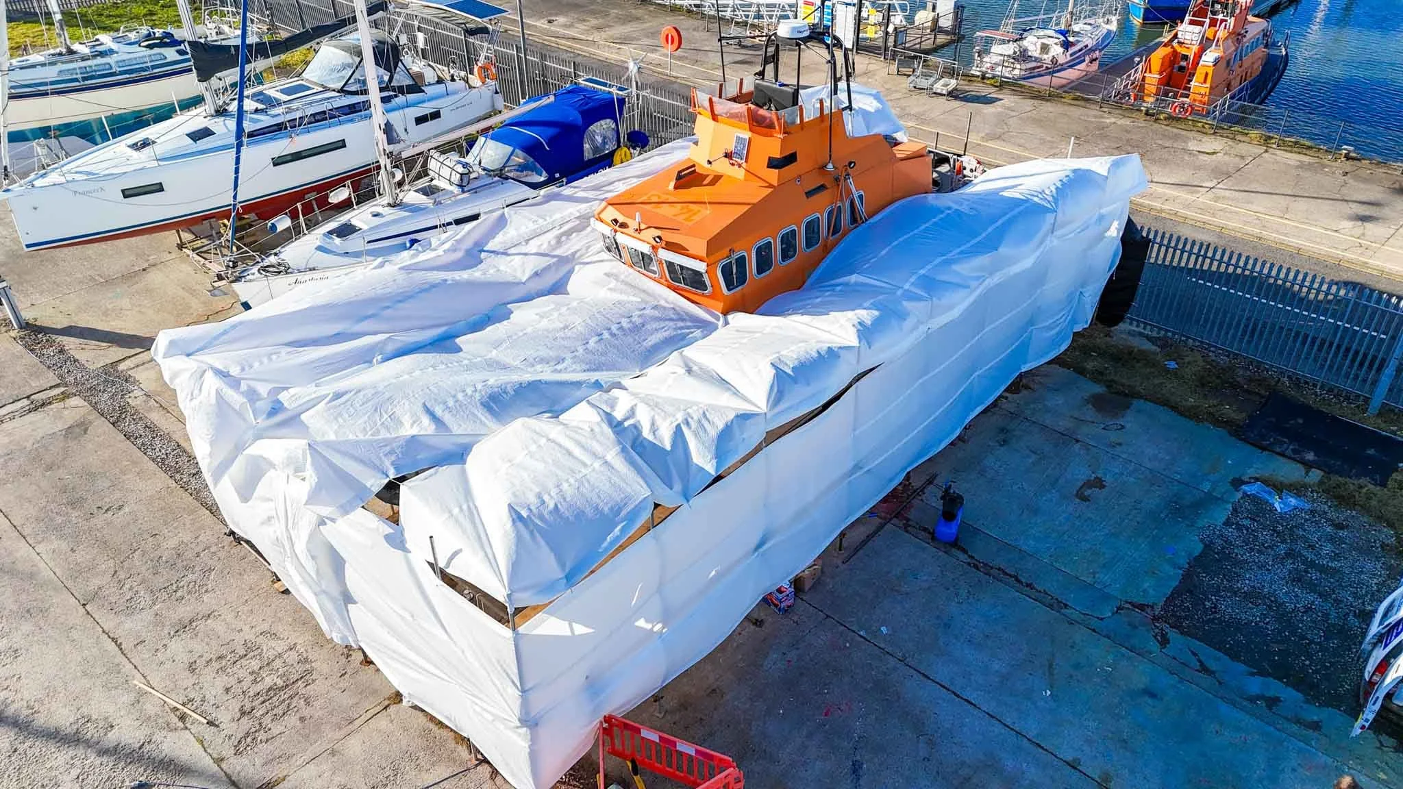 An orange boat is covered with white protective cloth in a boatyard, surrounded by other boats and sailboats on water, with a fence and marina in the background.