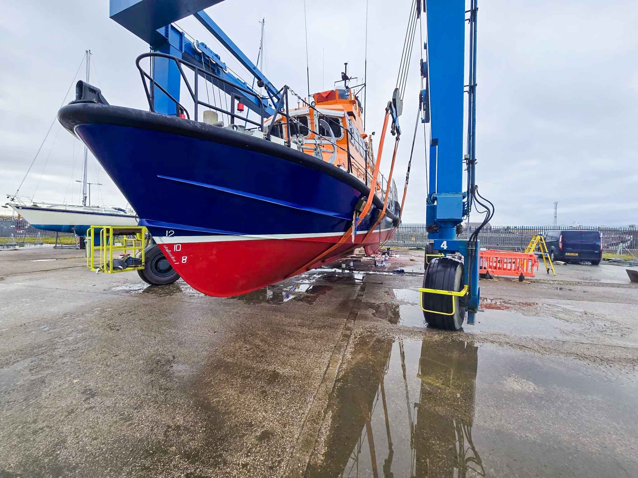 A boat being lifted out of the water on a crane at a boatyard, with the boat's hull reflected in puddles on the ground.