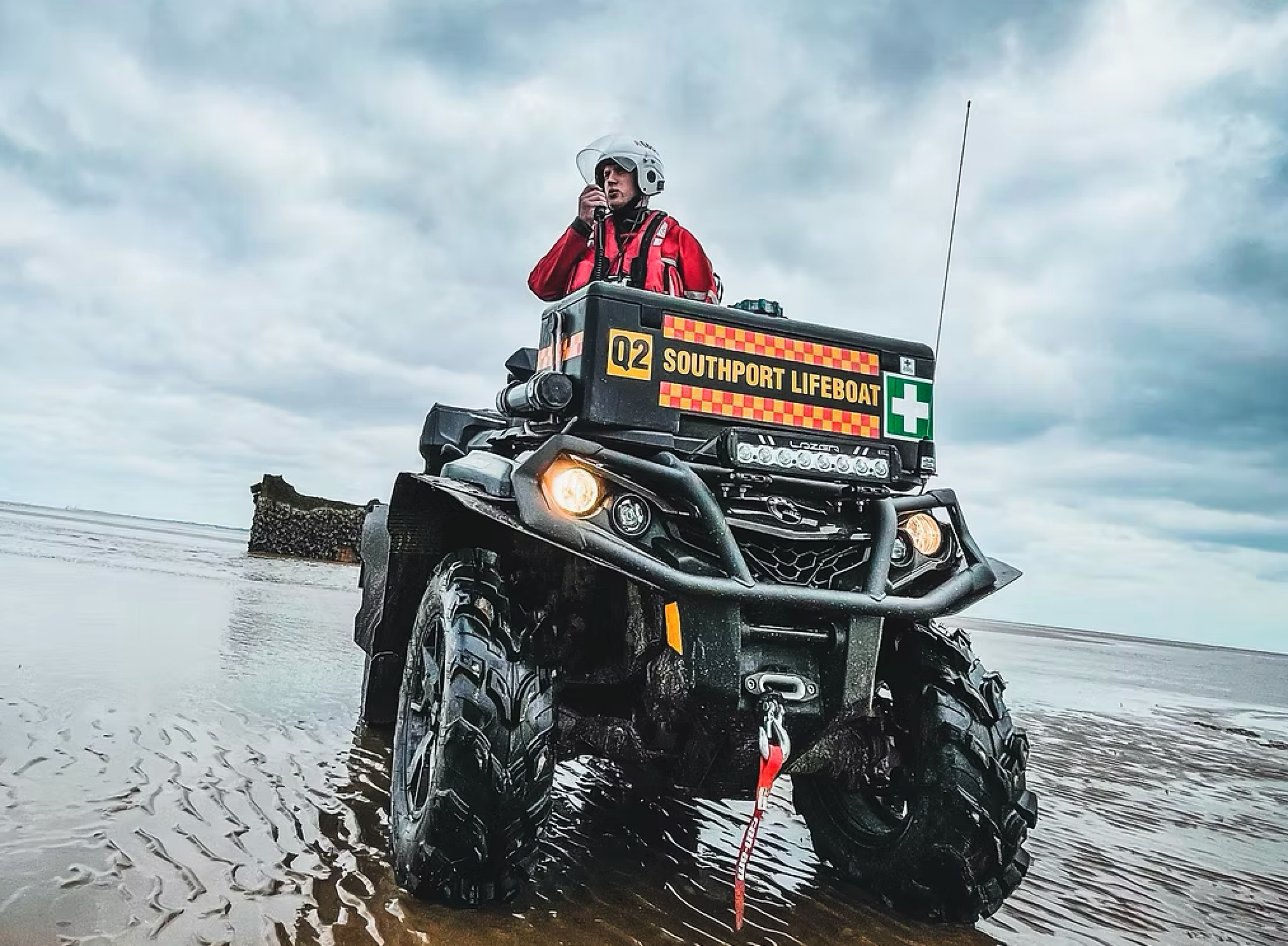 Emergency responder in red jacket and white helmet on a black all-terrain vehicle (ATV) in shallow water on a beach, with cloudy sky and a lifeguard sign on the ATV