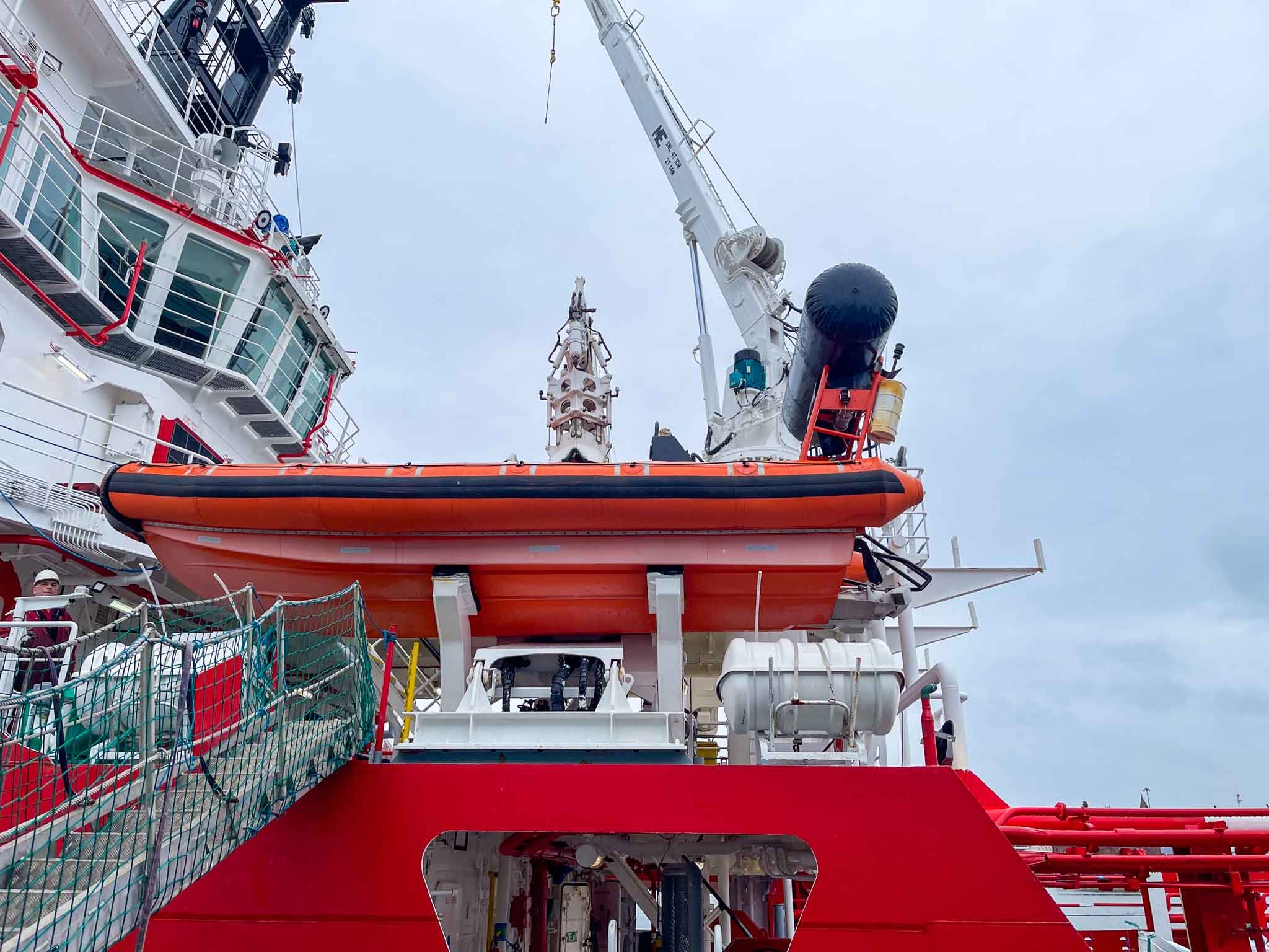 A close-up of the deck of a large vessel featuring safety equipment such as an orange rescue boat, a crane, pipes, and part of the ship's superstructure with windows and railings in the background.