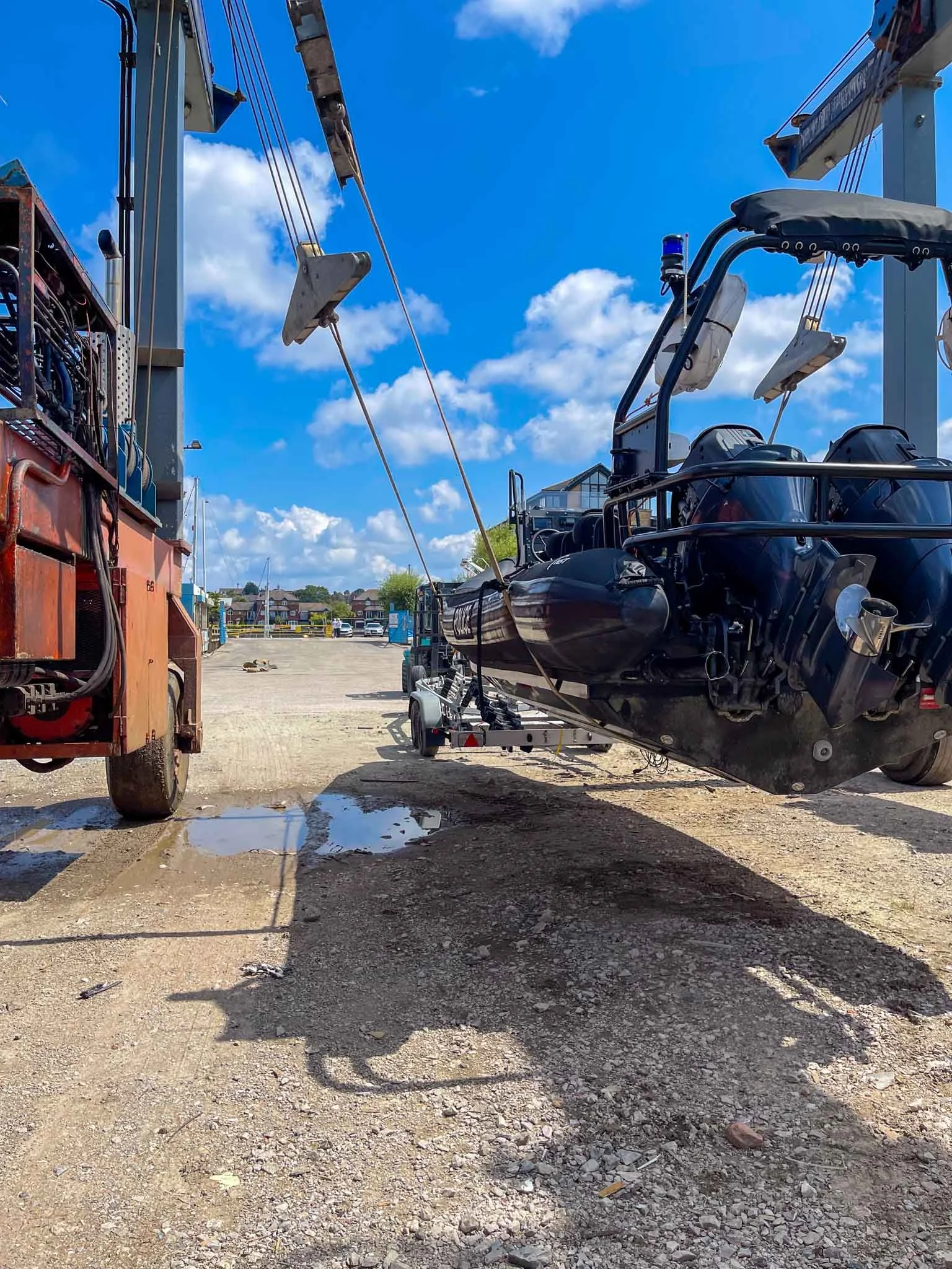 A boat is being lifted by a crane on a trailer, with blue skies and fluffy clouds overhead, and a small puddle on the gravel ground.