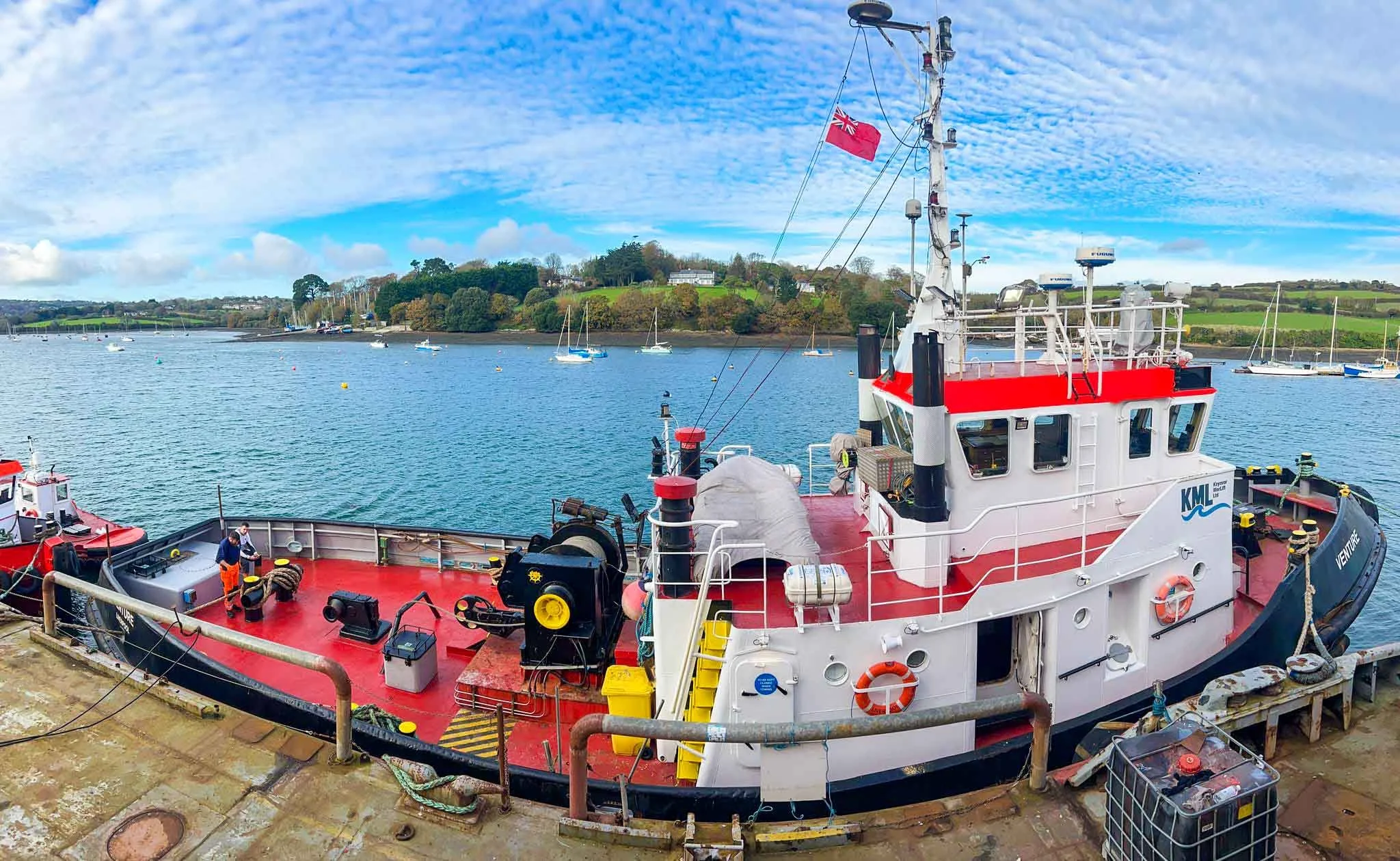 A red and white boat docked at a harbor, with a blue water body and sailboats in the background. There is an island with trees and grass across the water, and a partly cloudy sky above.
