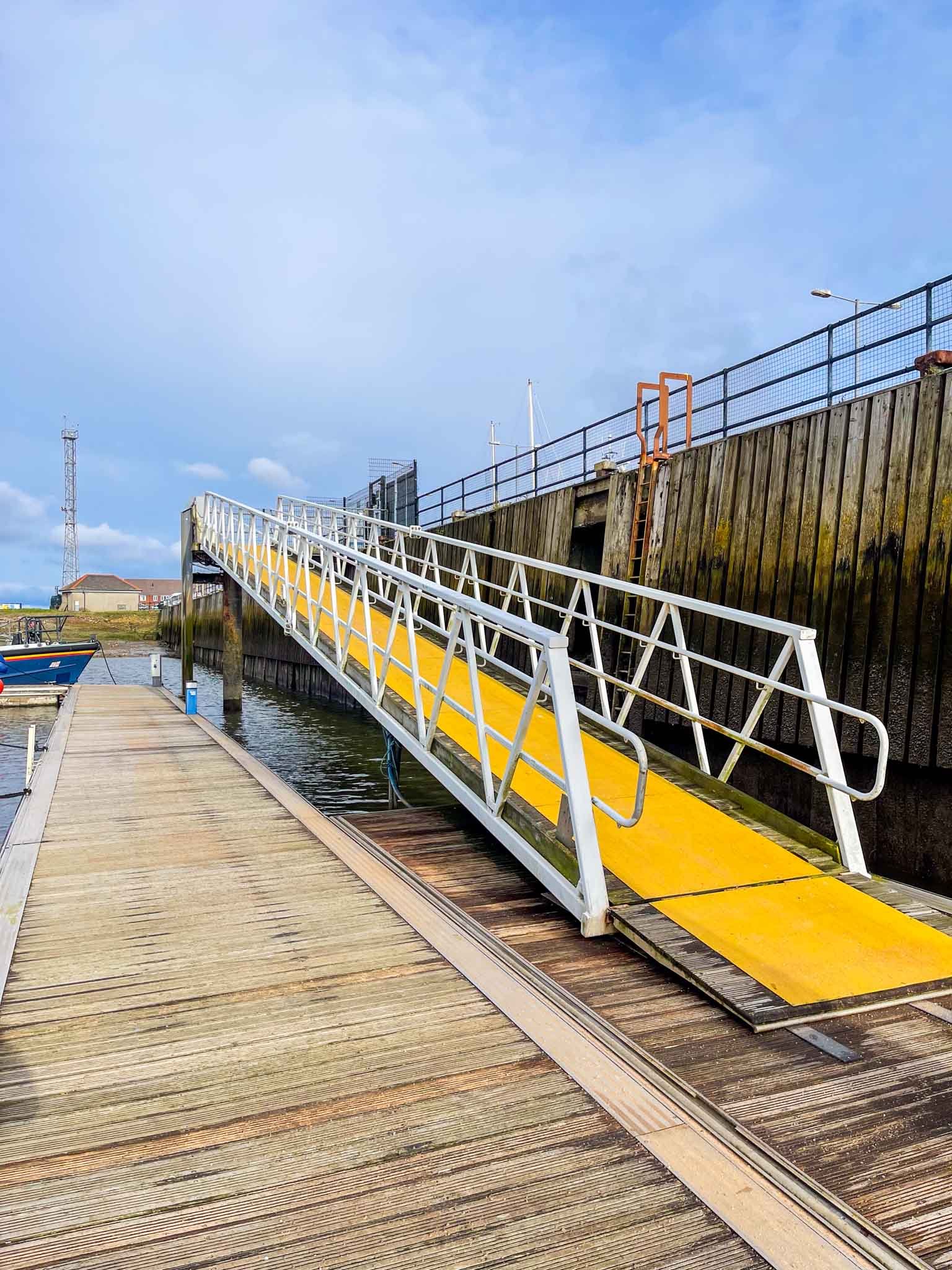 A yellow ramp leading up to a dock at a marina, with boats in the water and a cloudy sky above.