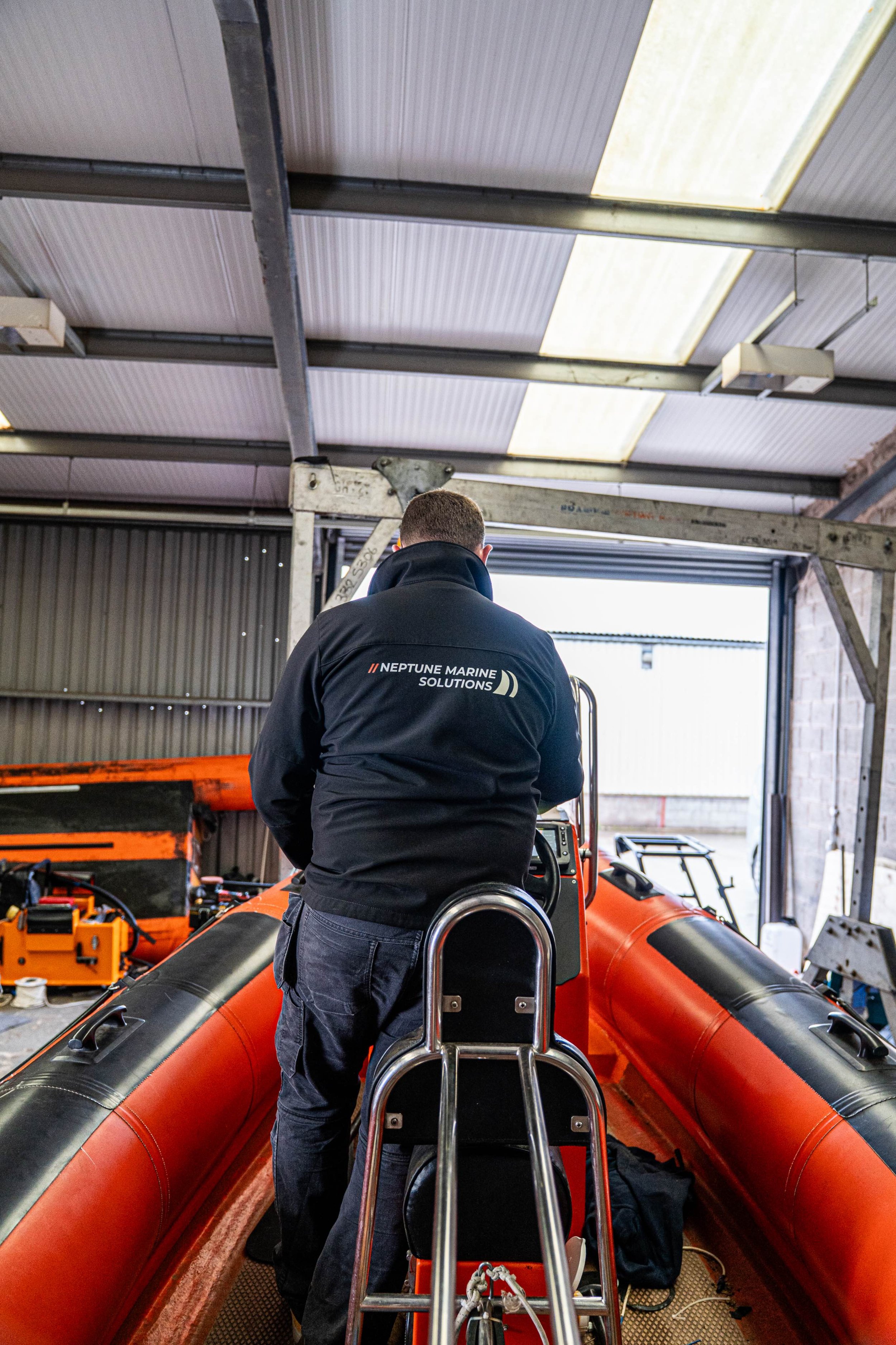 A man in a black jacket with 'Neptune Marine Solutions' on the back works on a small orange inflatable boat inside a workshop or garage.