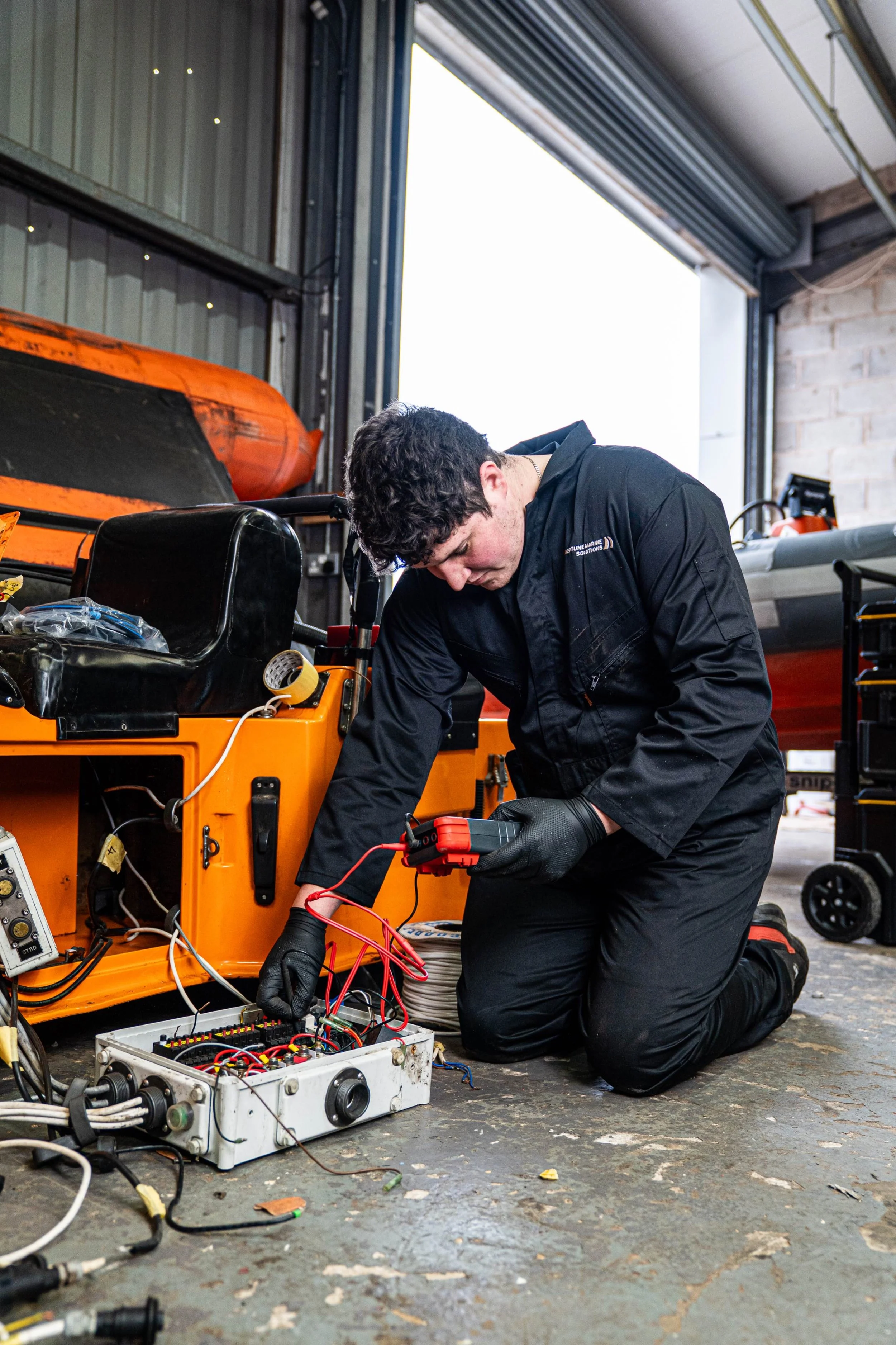 A mechanic working on an electrical system in a garage, kneeling on the floor and using a multimeter.