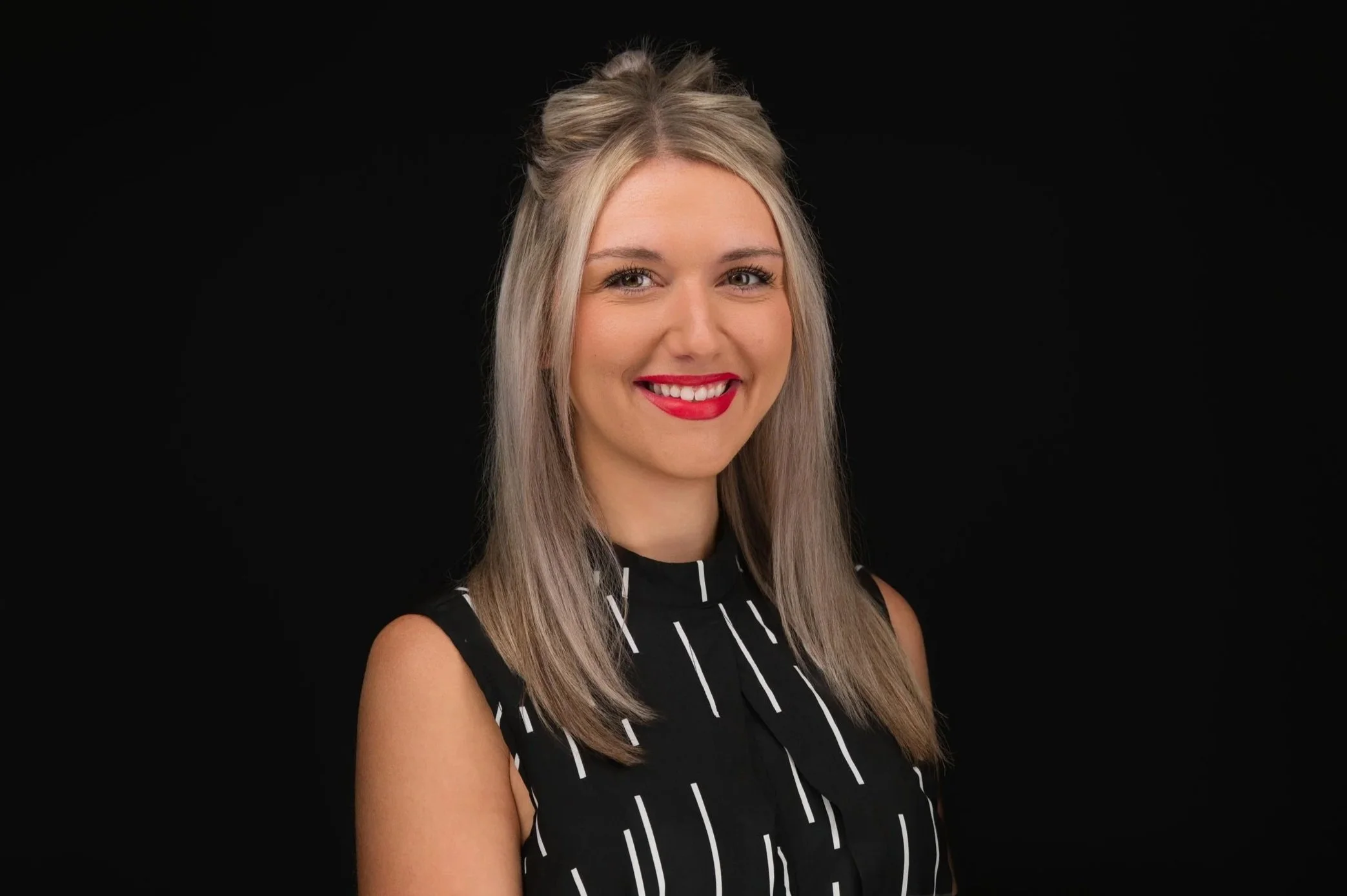 Portrait of a young woman with blonde hair styled in loose waves, wearing a black sleeveless top with white vertical lines, smiling against a black background.