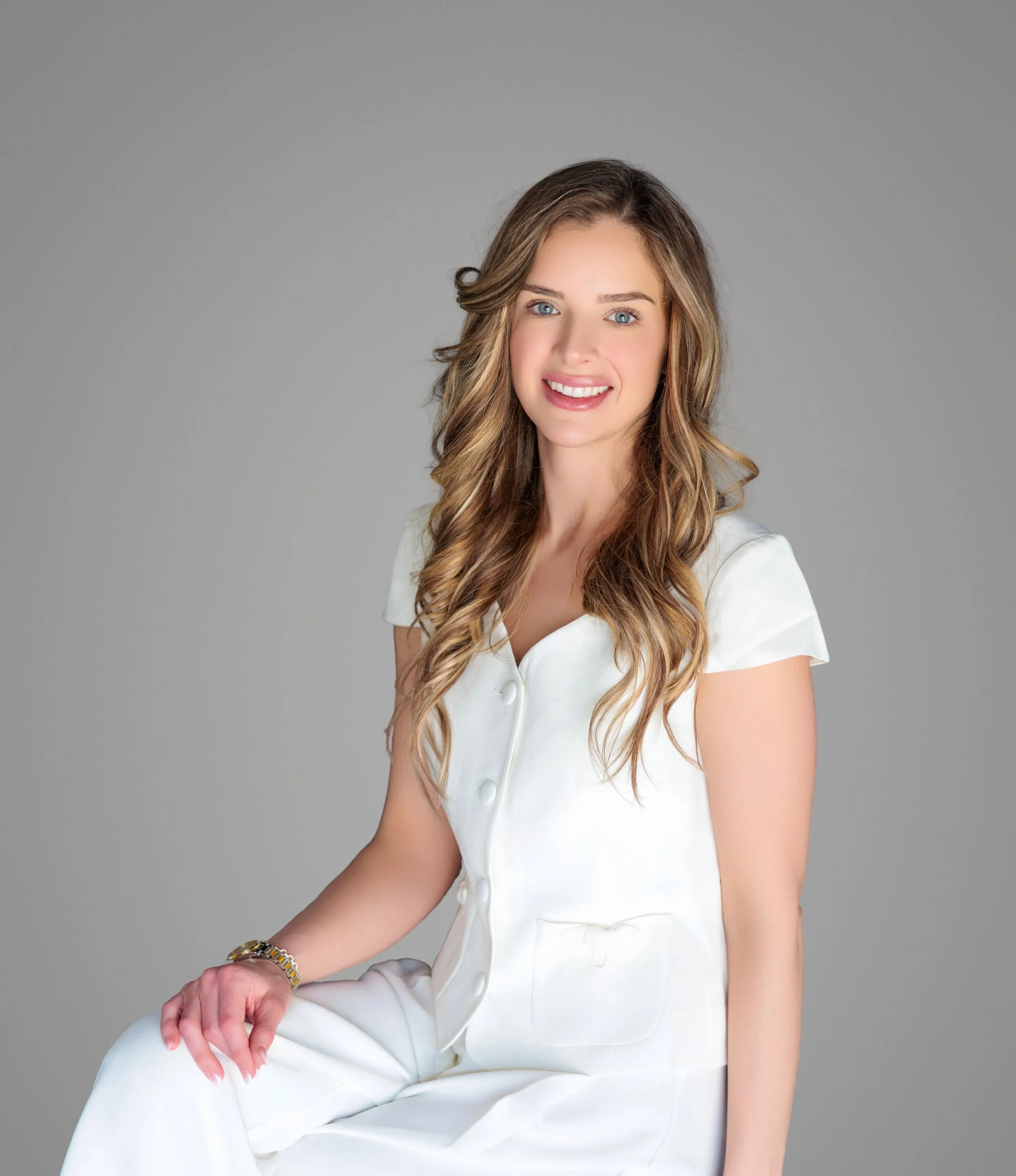 Young woman with long wavy brown hair wearing a white dress and sitting against a plain gray background.
