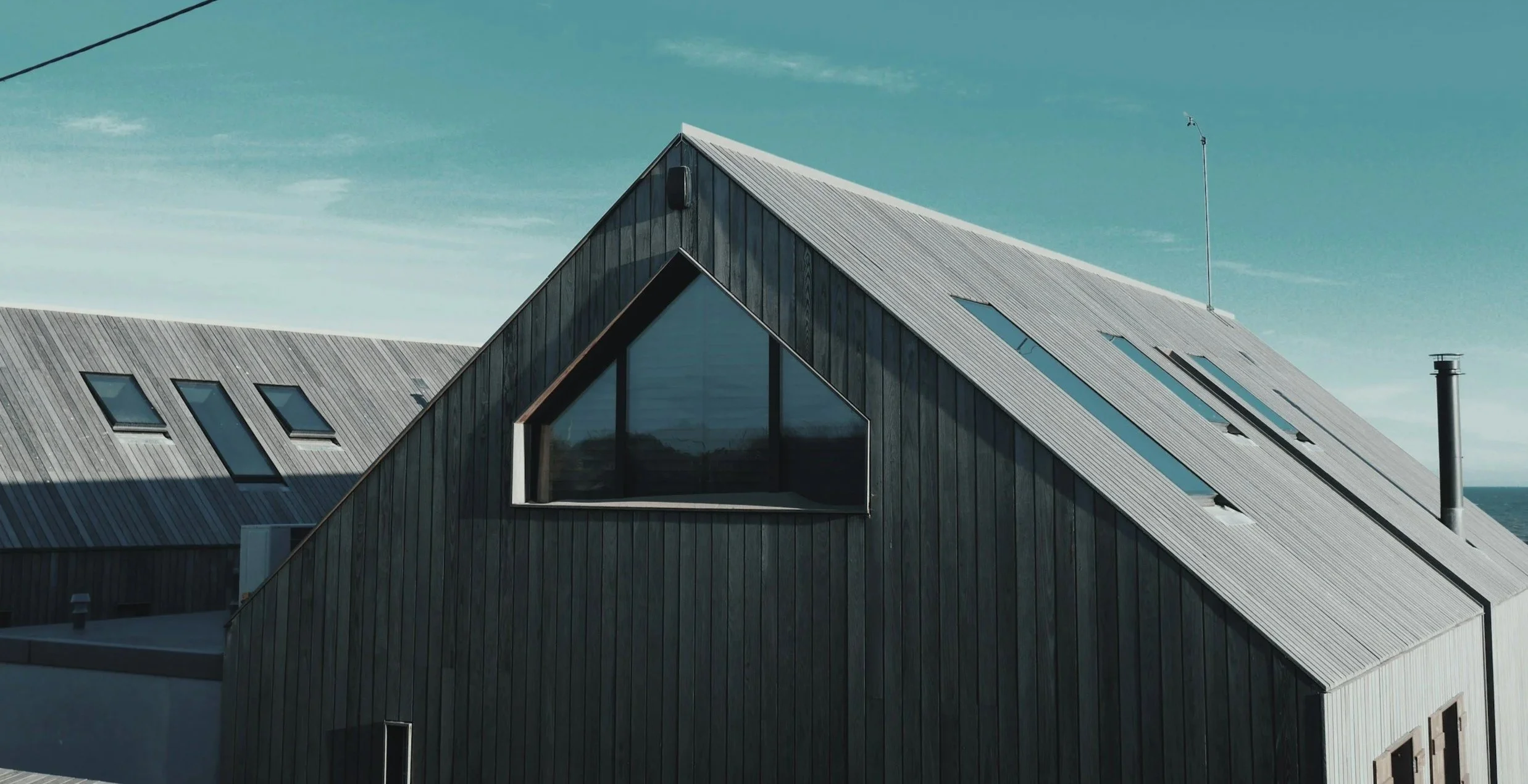 Modern house with dark wooden exterior, large triangular window, and skylights on metal roof, under blue sky.