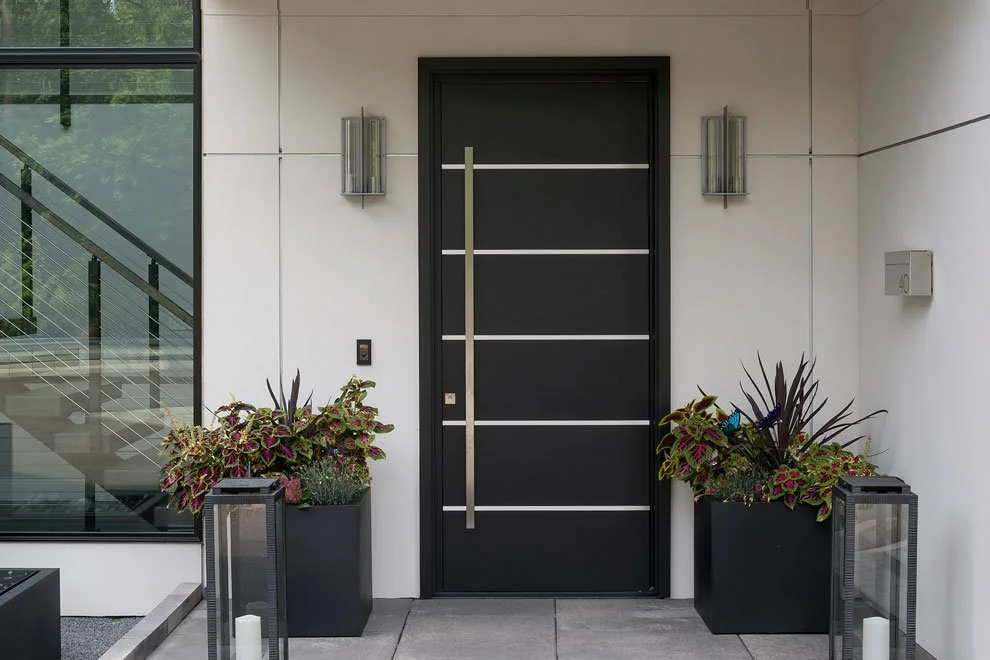 Modern black front door with sleek horizontal metal accents, flanked by two tall black planters filled with decorative plants, on a light-colored house exterior. Windows with a staircase visible inside are on the left side.
