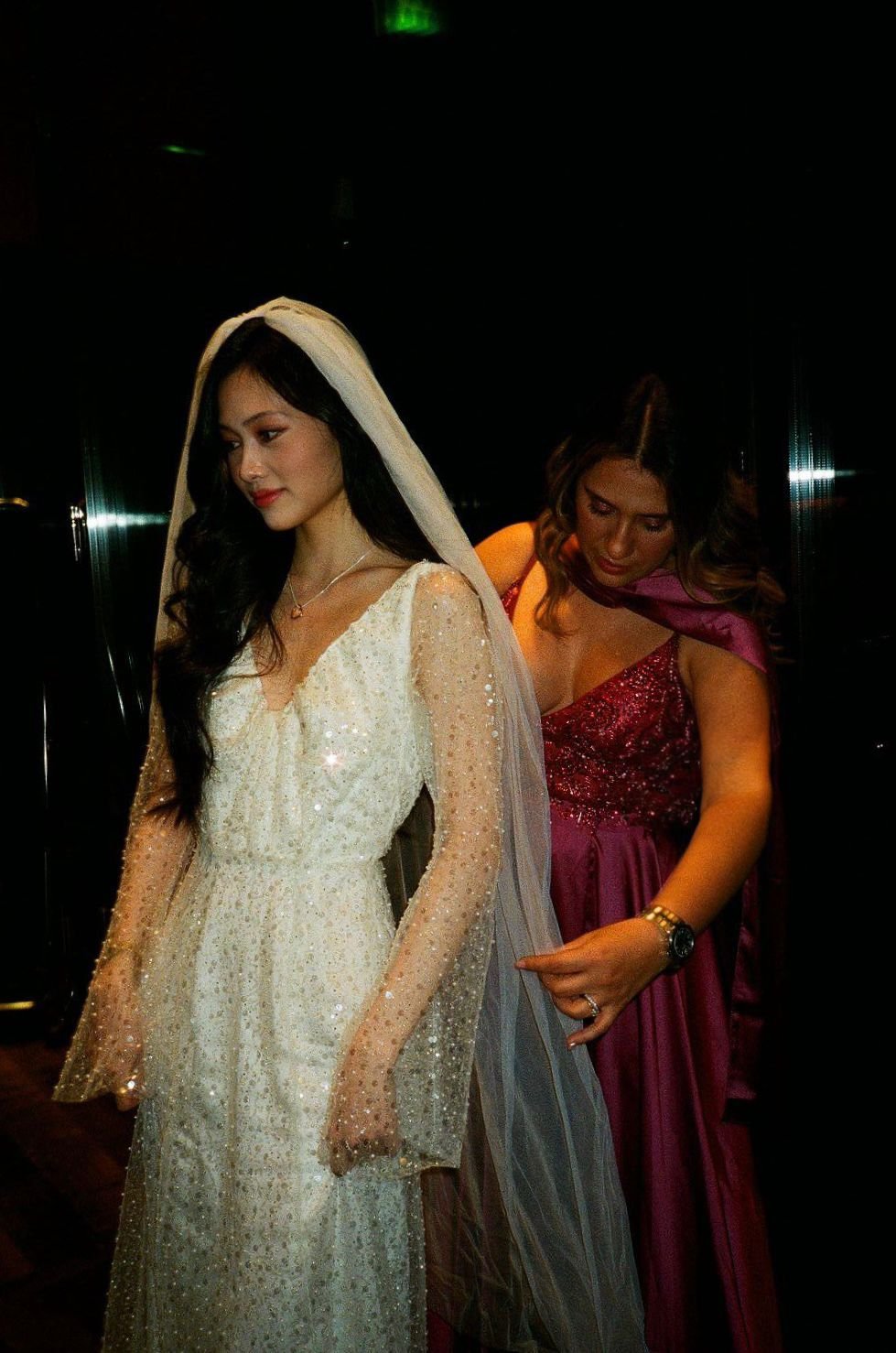 A woman dressed in a white, sparkling wedding gown with a veil is getting help adjusting her dress from another woman wearing a pink, sequined dress. The scene appears to be in a dimly lit room.