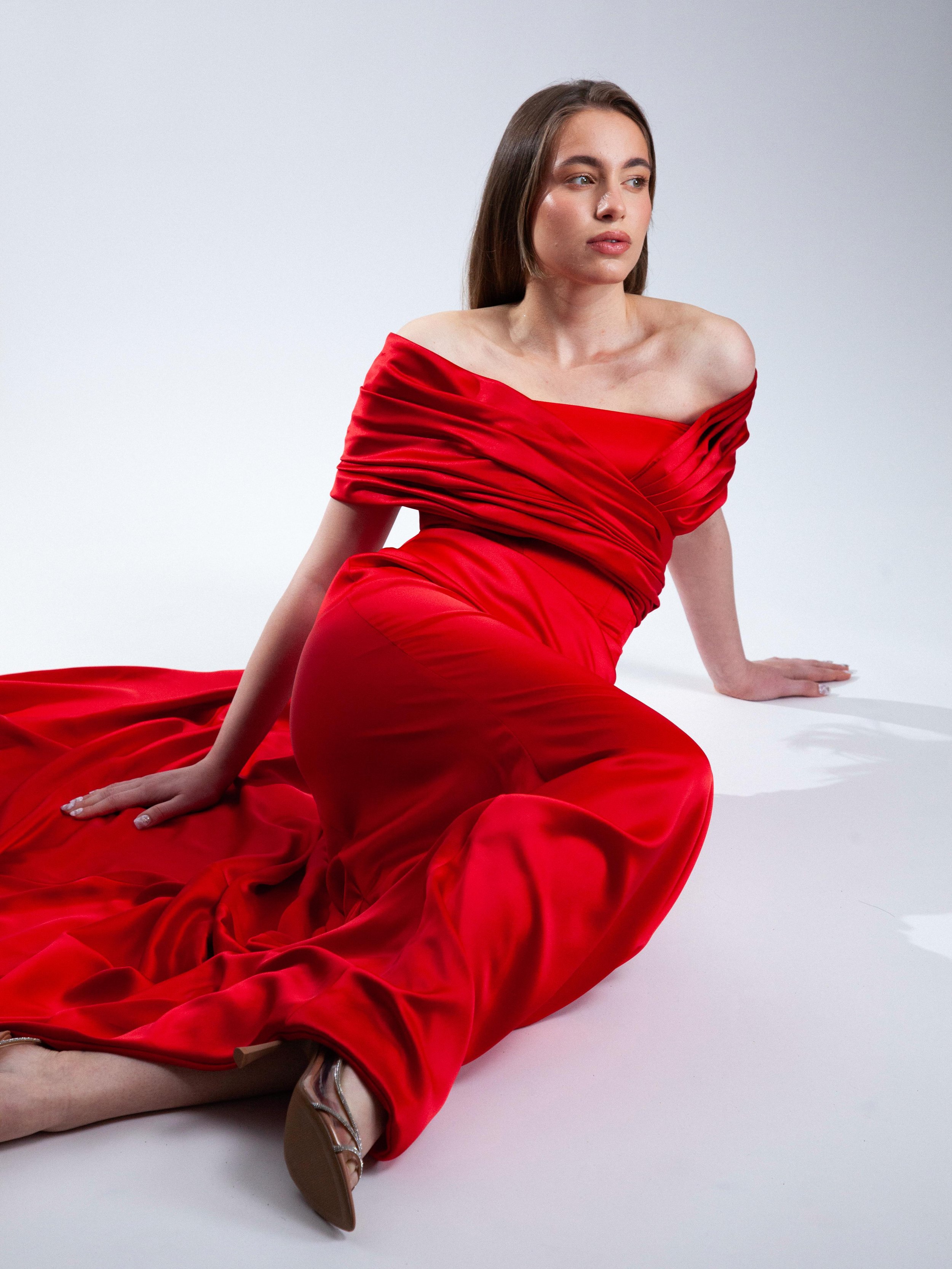 A woman in a red off-the-shoulder gown sitting on the floor against a plain white background.