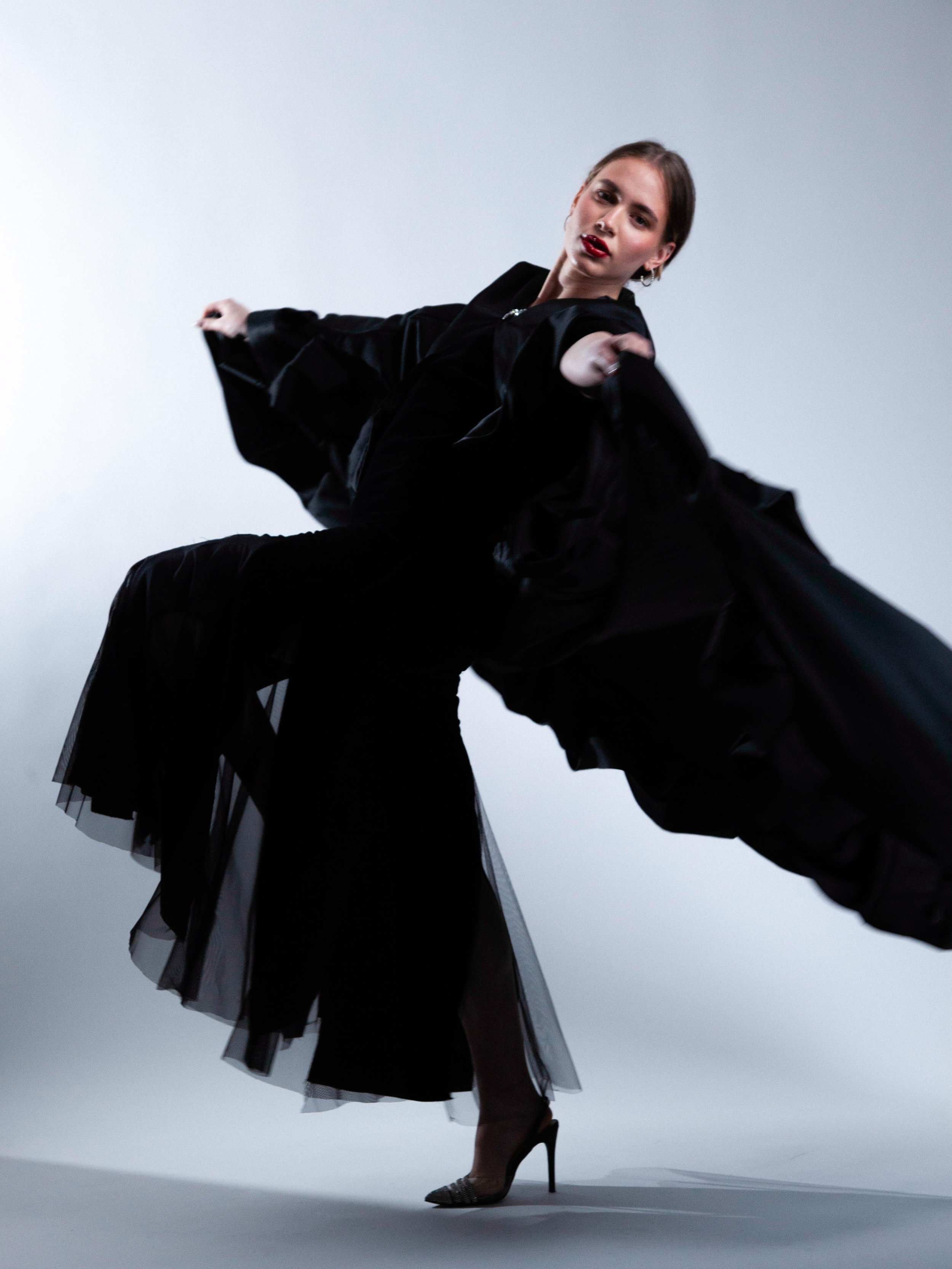 A woman in a black, flowing dress with high heels is dancing against a plain background, captured from a low angle.