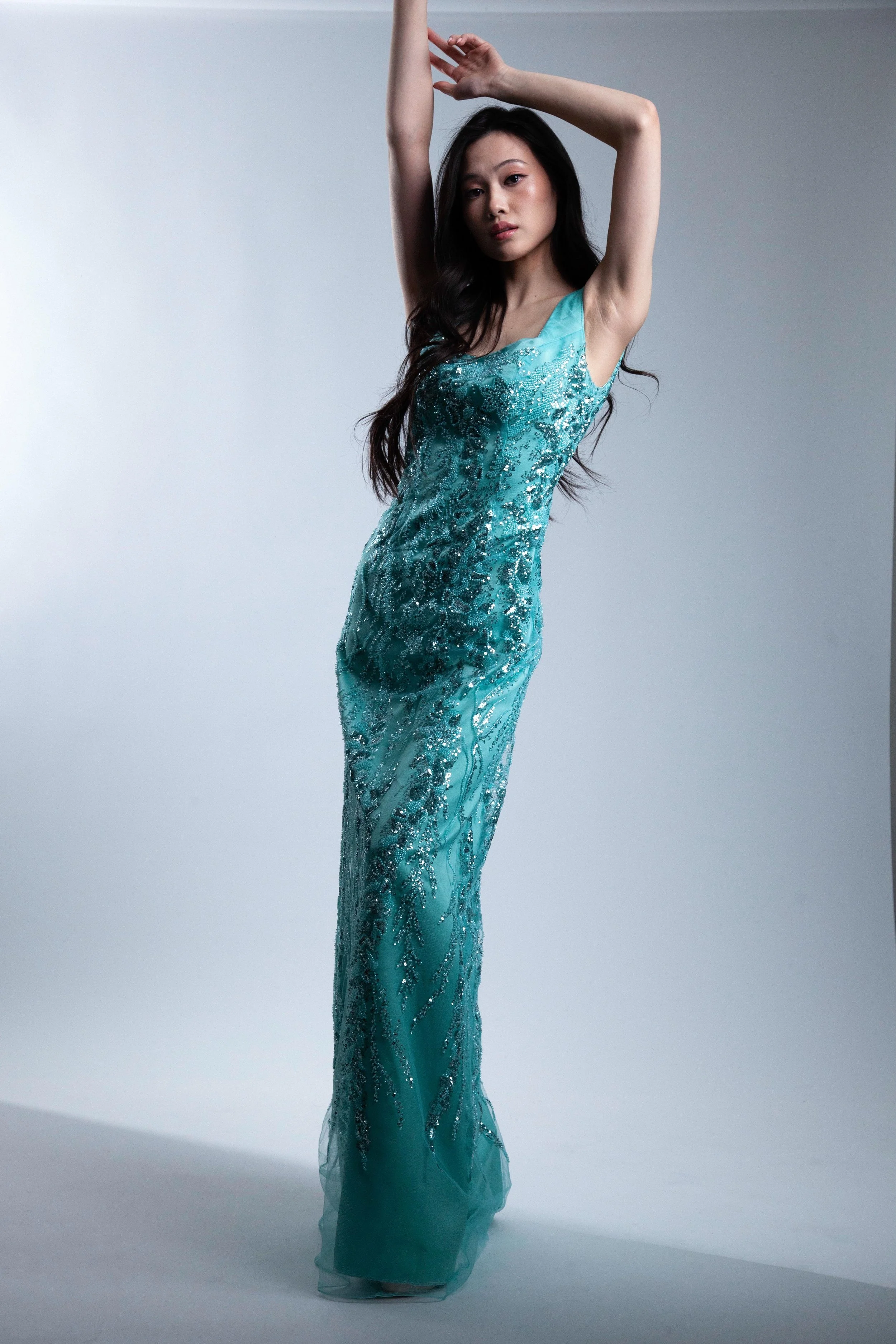 A woman in a long, shiny blue dress with silver embellishments, standing with arms raised above her head against a plain gray background.