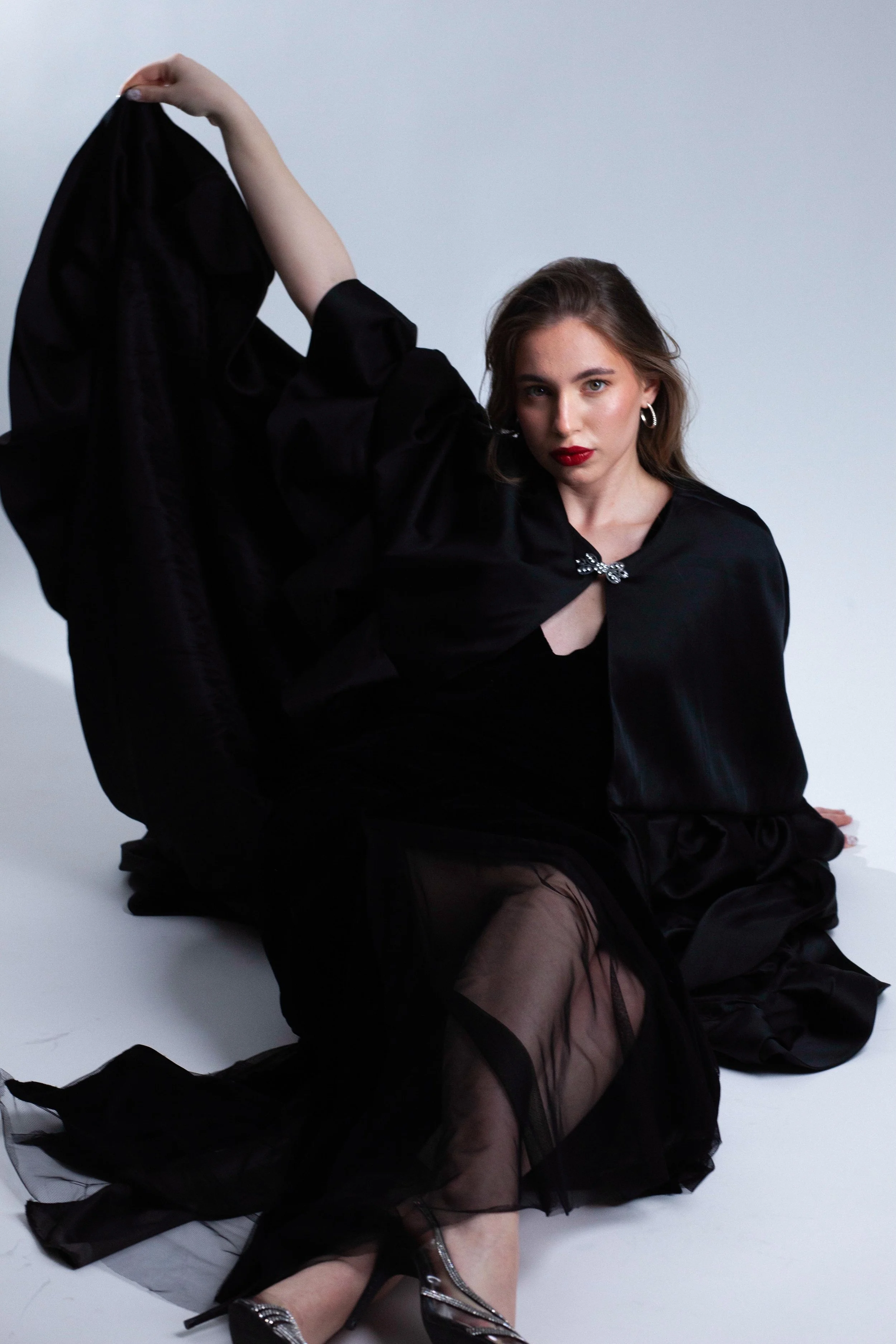 A woman with long hair and red lipstick, dressed in black, sitting on the floor with one leg extended and the other bent, wearing high heels and sheer black fabric, posing against a plain white background.