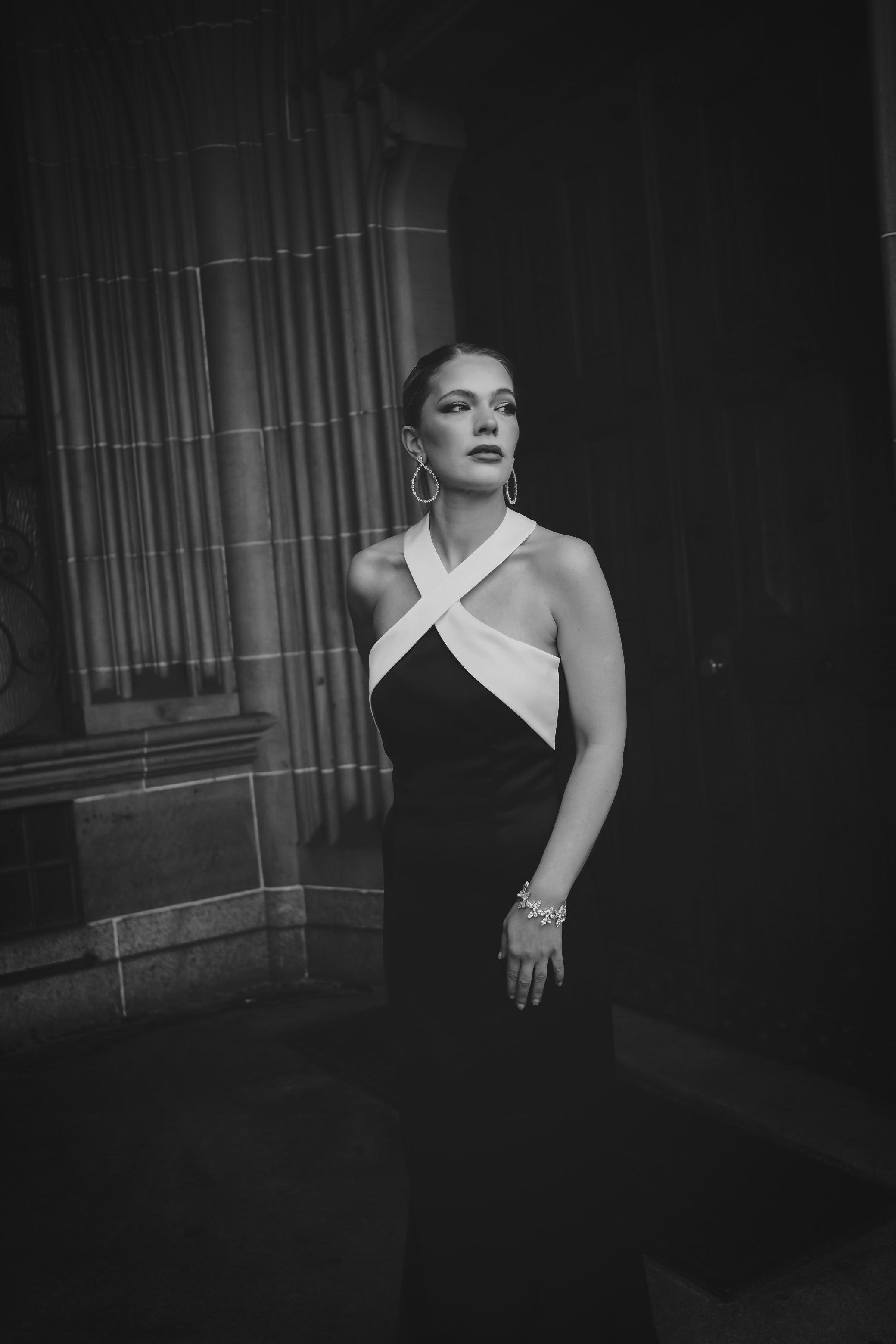 A woman in a formal black and white gown with halter neckline standing indoors near stone walls and heavy drapery, looking away from the camera.