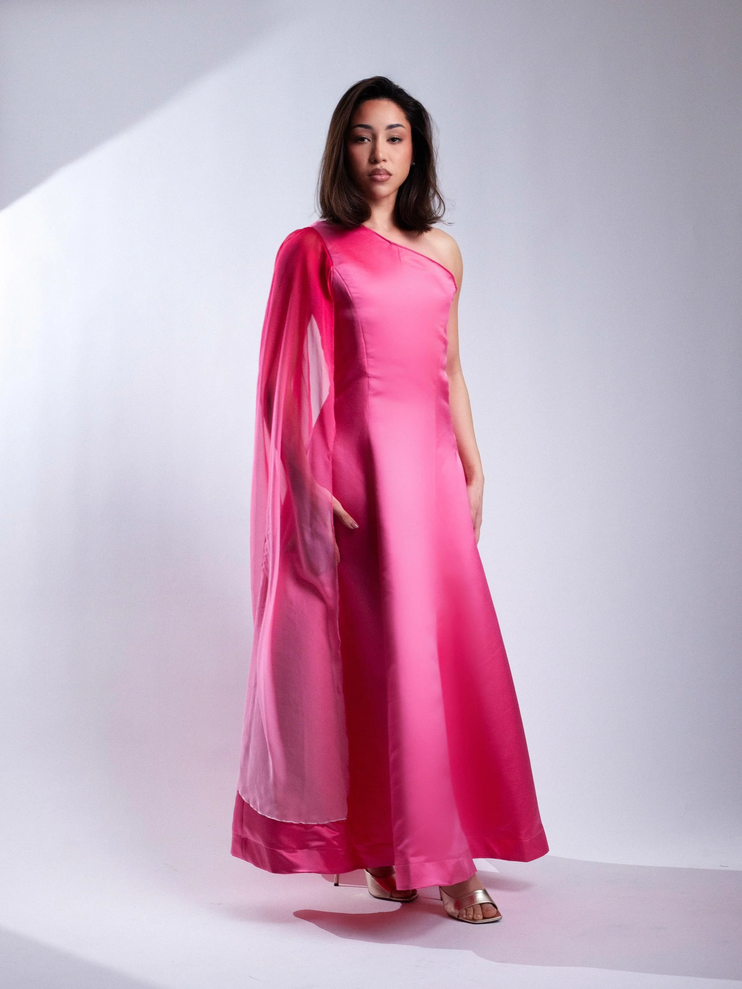 Woman wearing a pink one-shoulder gown with a matching sheer cape, standing on a white background.