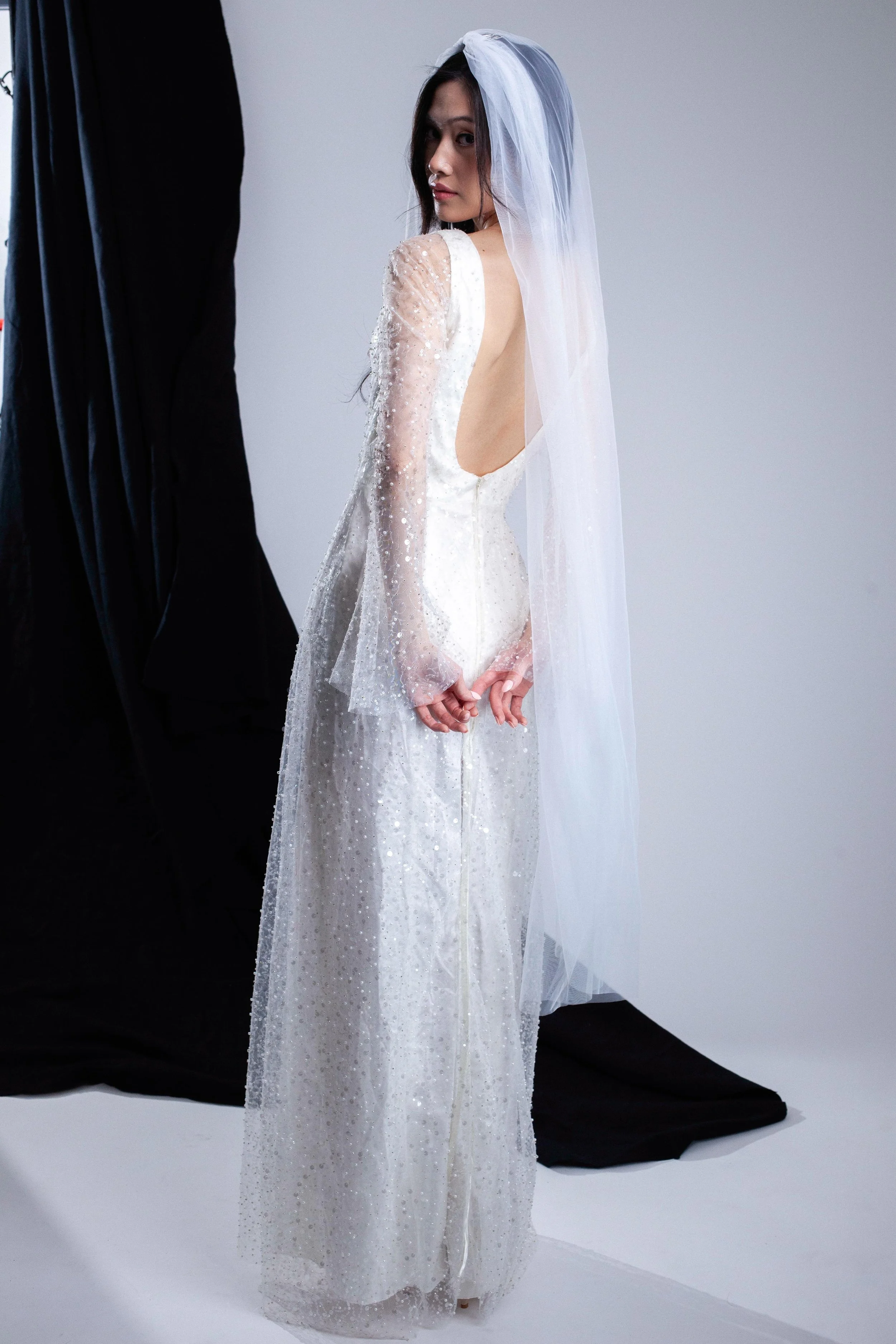 A woman wearing a white wedding dress with long sheer sleeves and a veil, standing against a plain background, facing slightly away but looking over her shoulder.