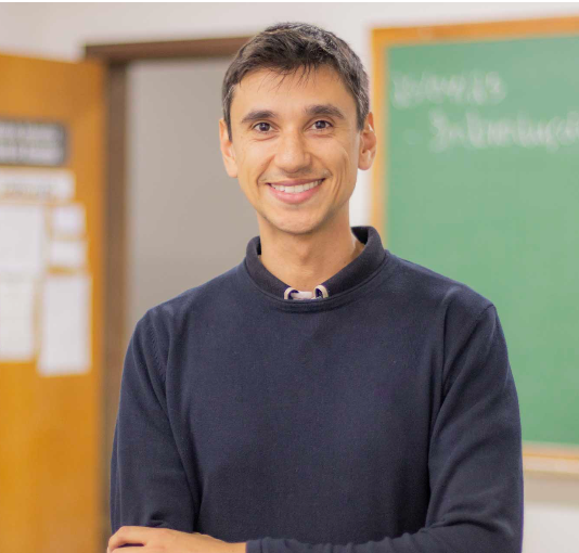 Homem sorrindo em sala de aula com quadro verde ao fundo