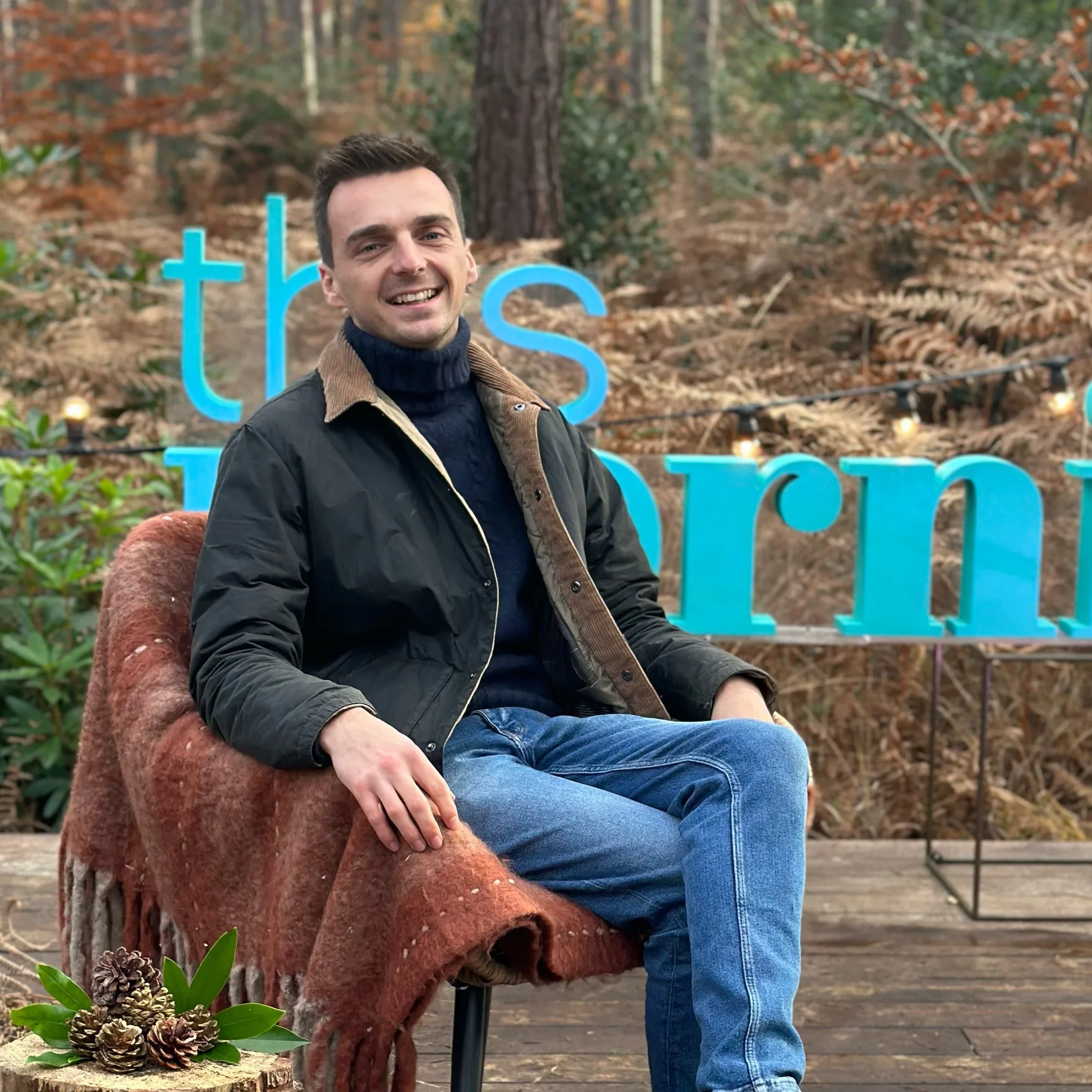 A young man sitting outdoors on a wooden deck with a brown blanket. Behind him, there is a blue sign with part of the word 'the' showing, and a forest with brown leaves and trees in the background.