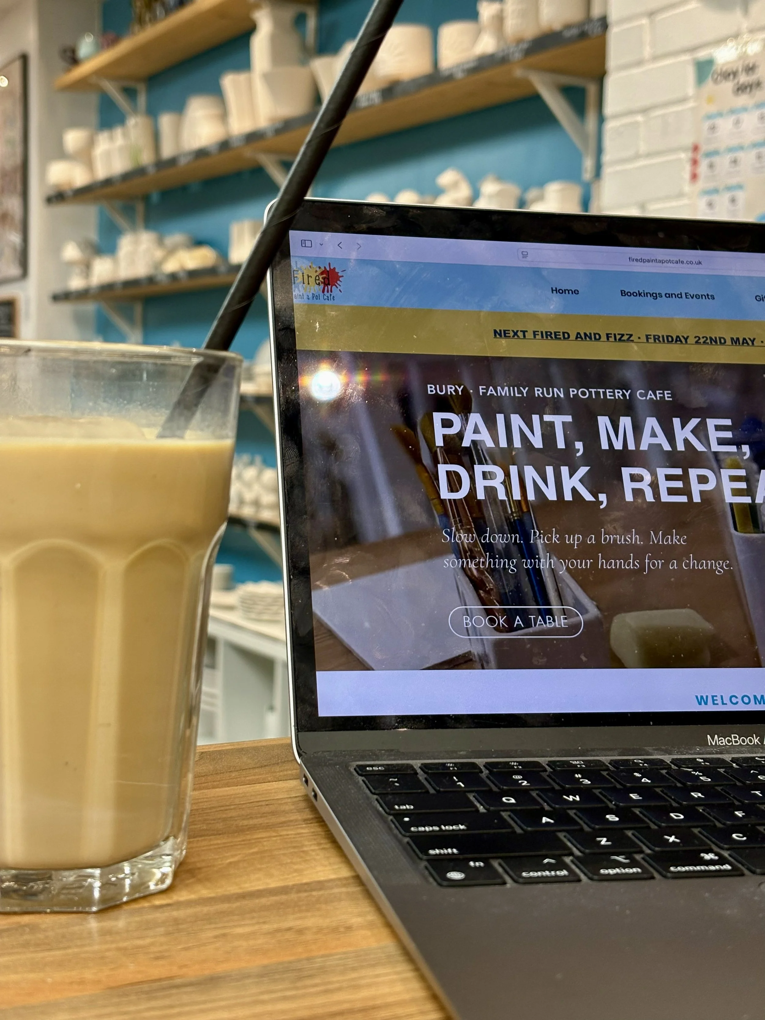 The redesigned Fired Paint a Pot Cafe website displayed on a laptop in the studio, with shelves of blank pottery behind and a coffee alongside.