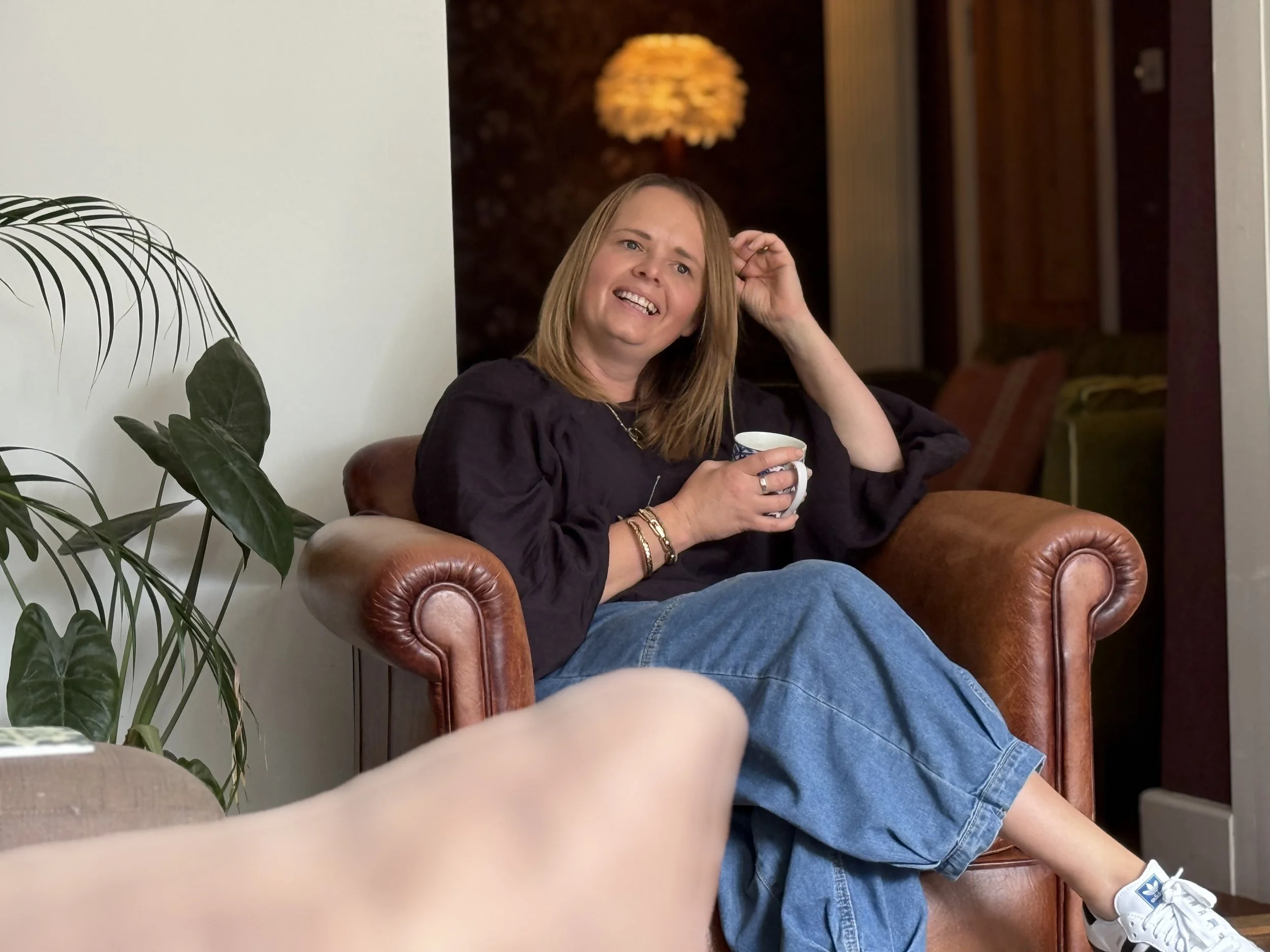 A woman with shoulder-length light brown hair sitting in a brown leather armchair, holding a white mug, smiling, and scratching her head. She is wearing a black top, blue jeans, white sneakers, and several bracelets.