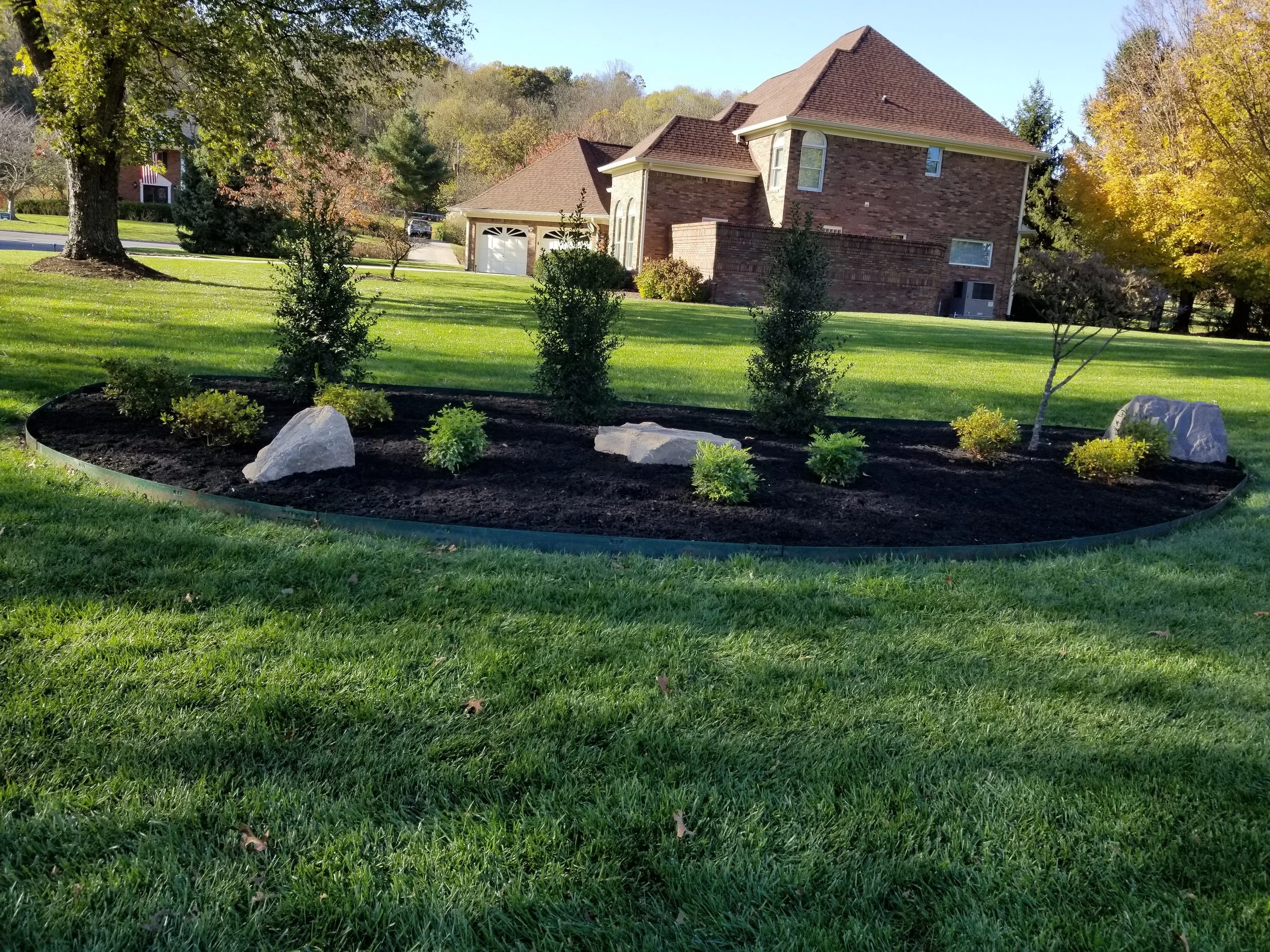 A landscaped yard with a flower bed bordered by a black plastic edge, containing small bushes, trees, and two large rocks, with a brick house in the background and a well-manicured lawn.
