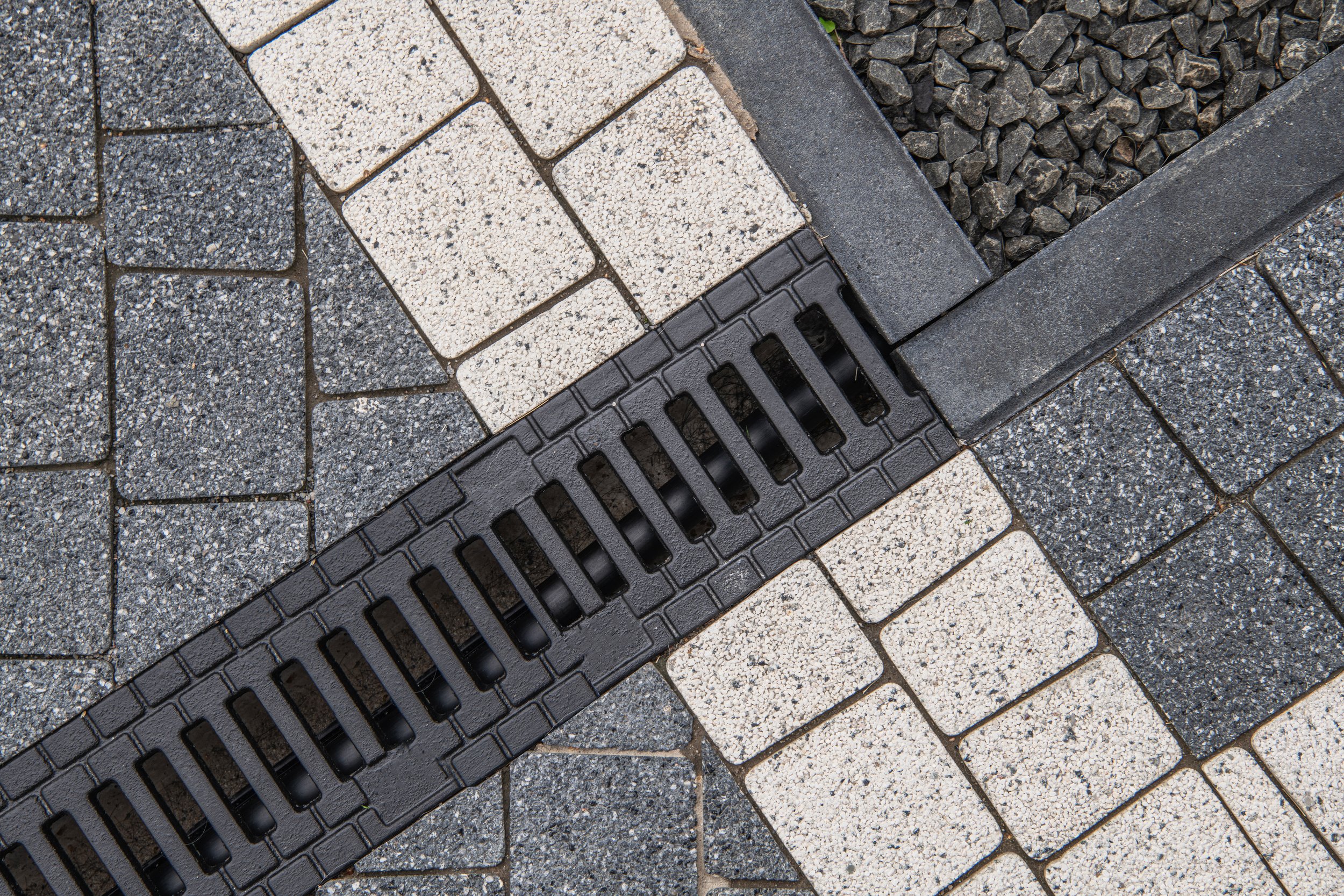 Close-up view of a black drainage grate on a paved surface with gray and white bricks and black gravel surrounding it.