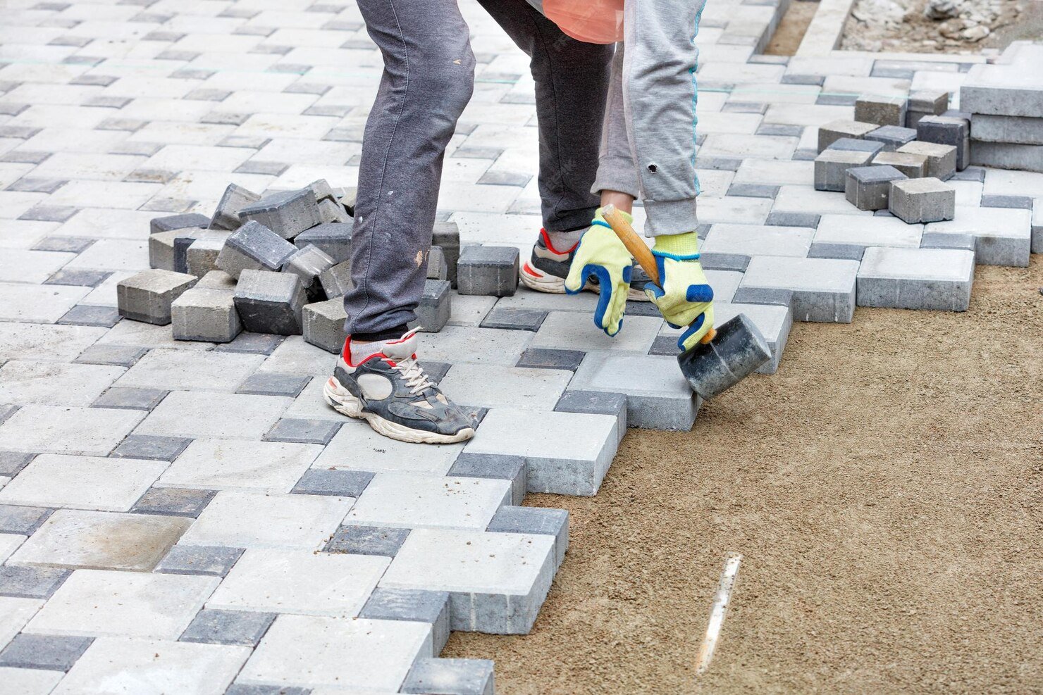Two workers laying concrete pavers on a sidewalk, one with a hammer and gloves, arranging grey and black paving stones over sand.