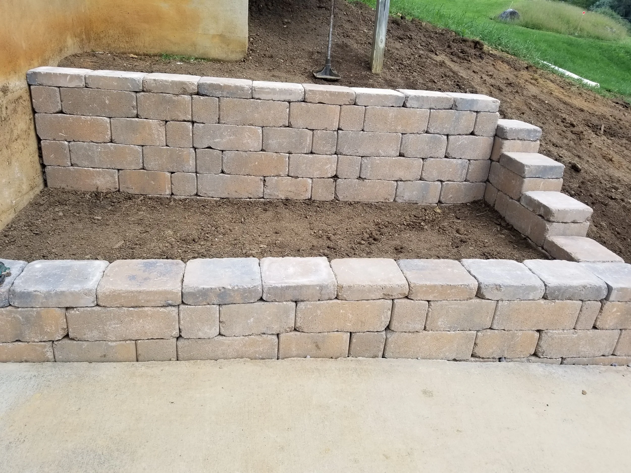 A brick retaining wall with two levels, a small set of three steps on the side, built on dirt with wood posts and a shovel nearby.