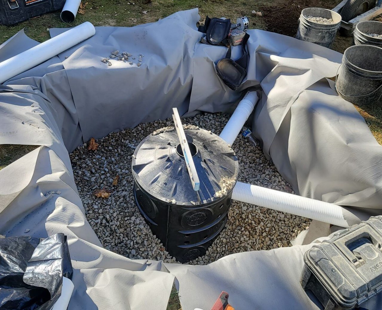 A construction site with a large round plastic tank in an excavation, surrounded by gravel and lined with gray fabric. Piping extends from the tank, and there are various tools, buckets, and construction materials around the area.