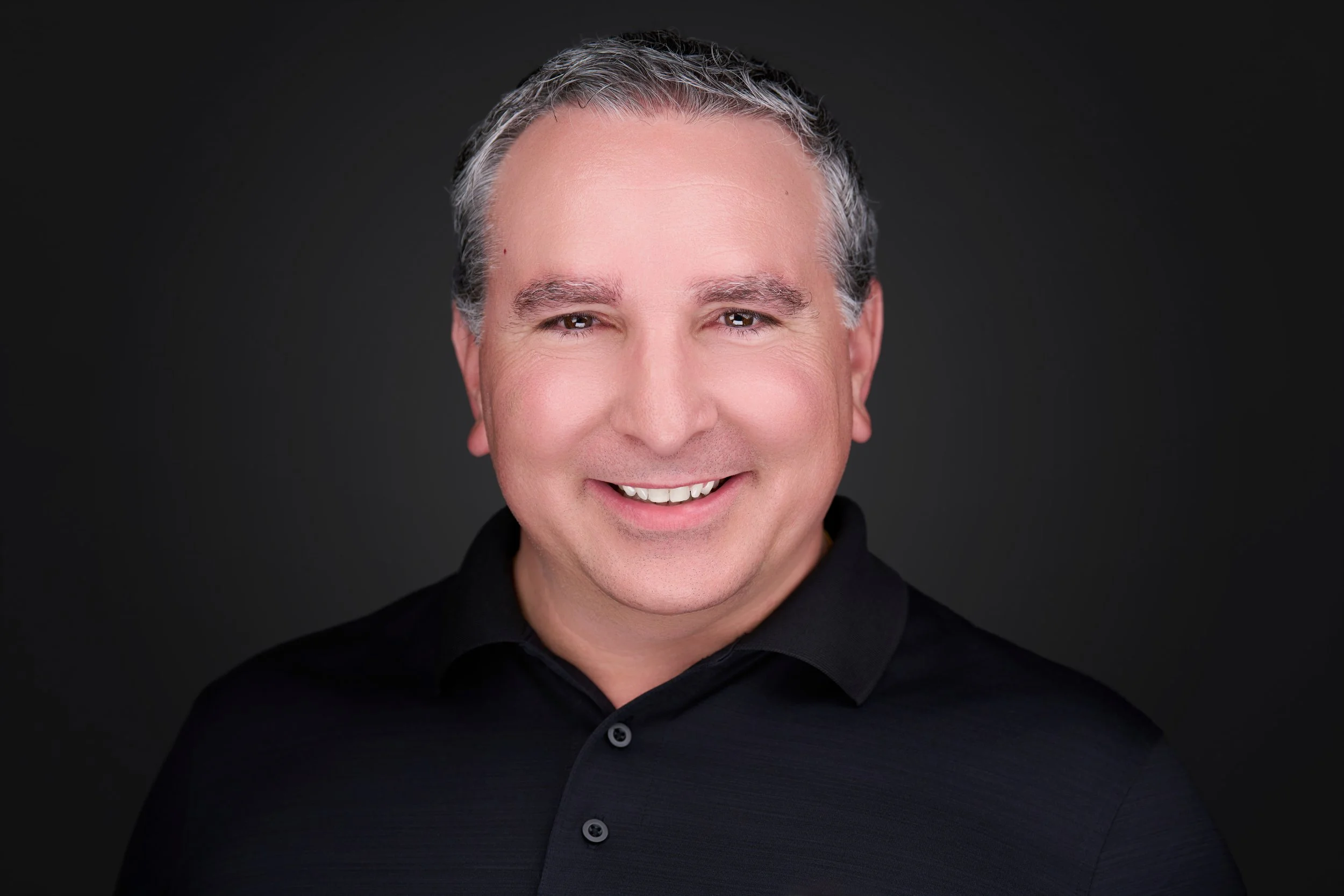 Close-up portrait of a smiling middle-aged man with gray hair, wearing a black collared shirt, against a plain dark background.