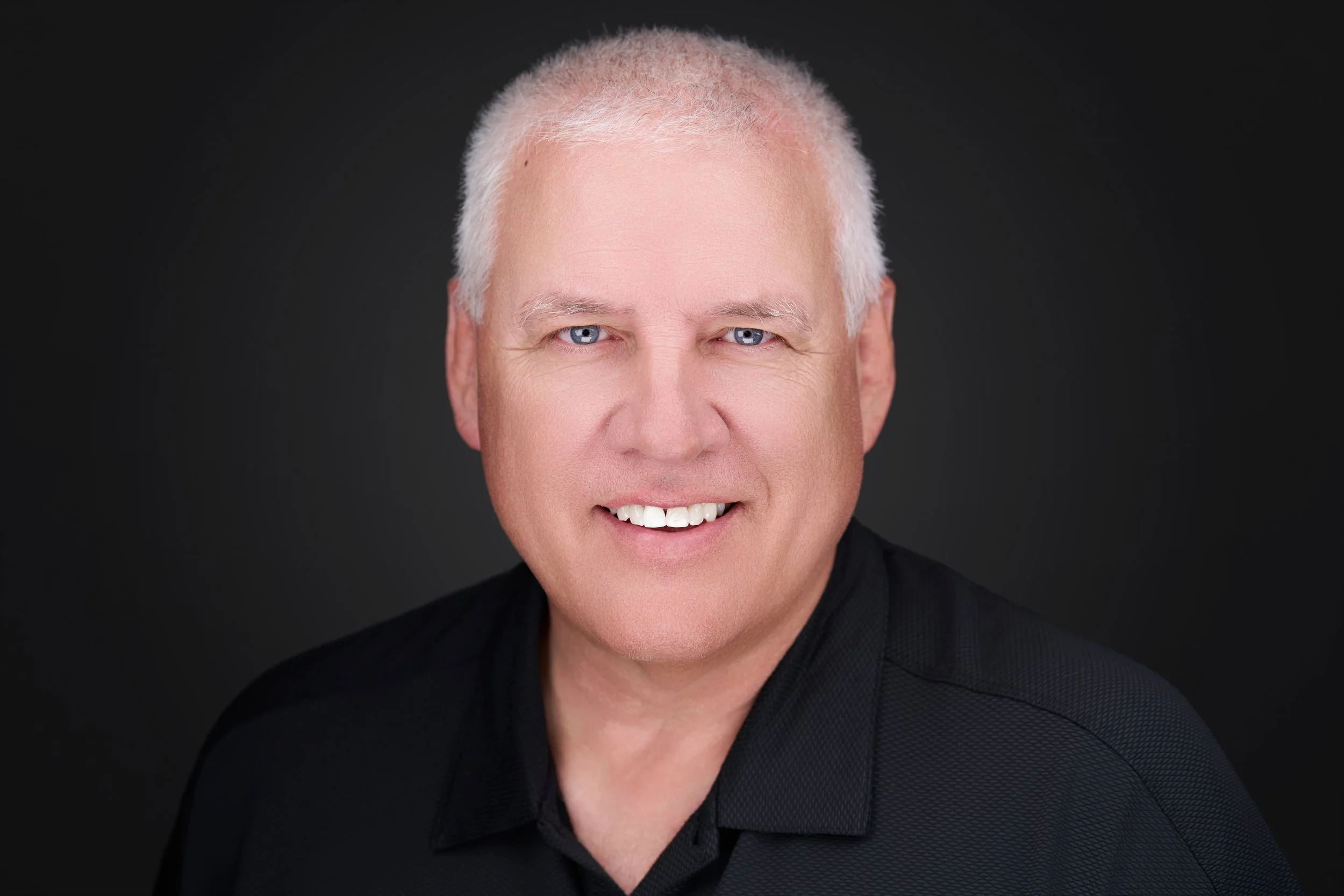 Close-up of a smiling older man with short white hair and blue eyes, wearing a black shirt, against a dark background.
