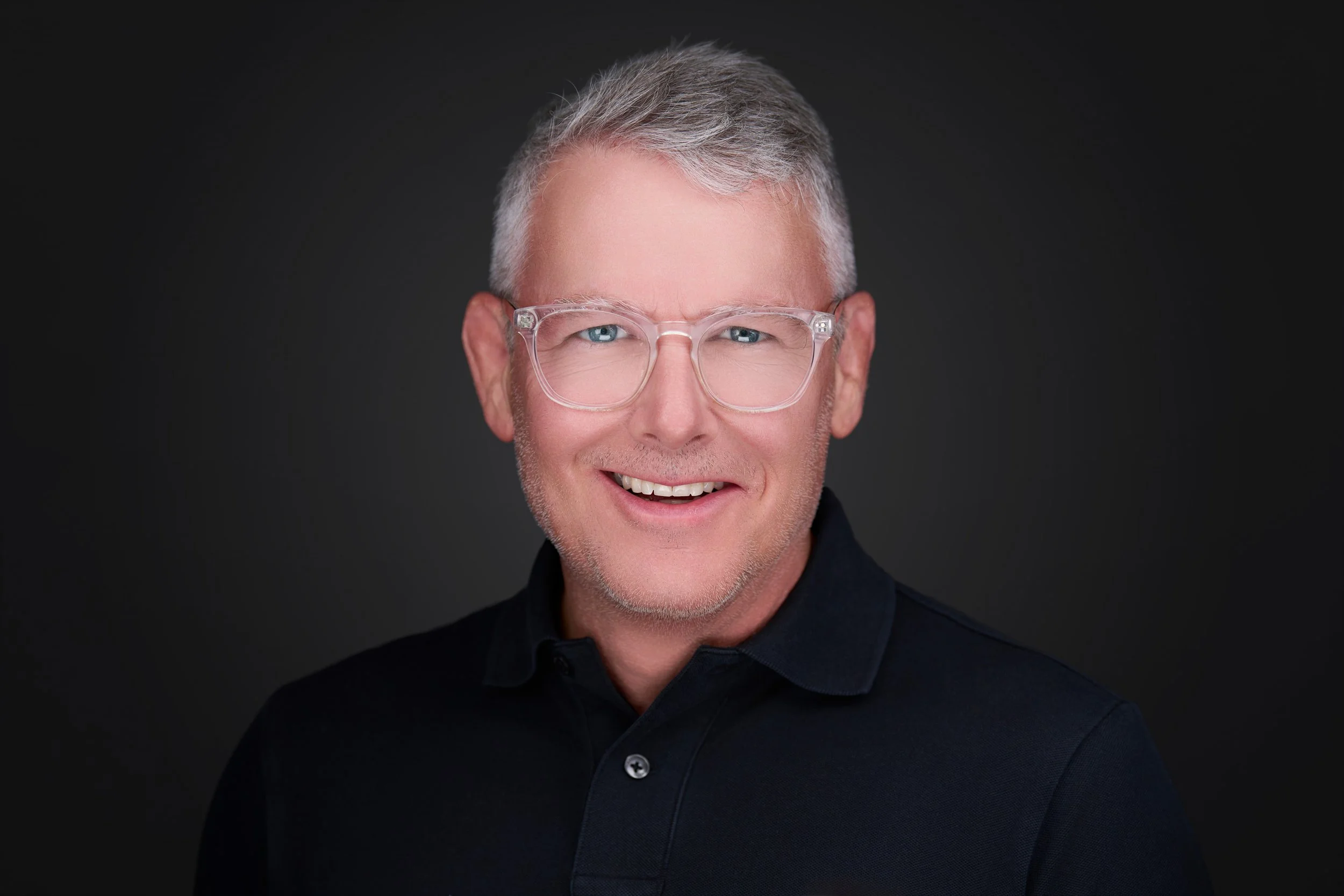 Close-up portrait of a smiling middle-aged man with gray hair and blue eyes, wearing clear eyeglasses and a black collared shirt, against a dark background.