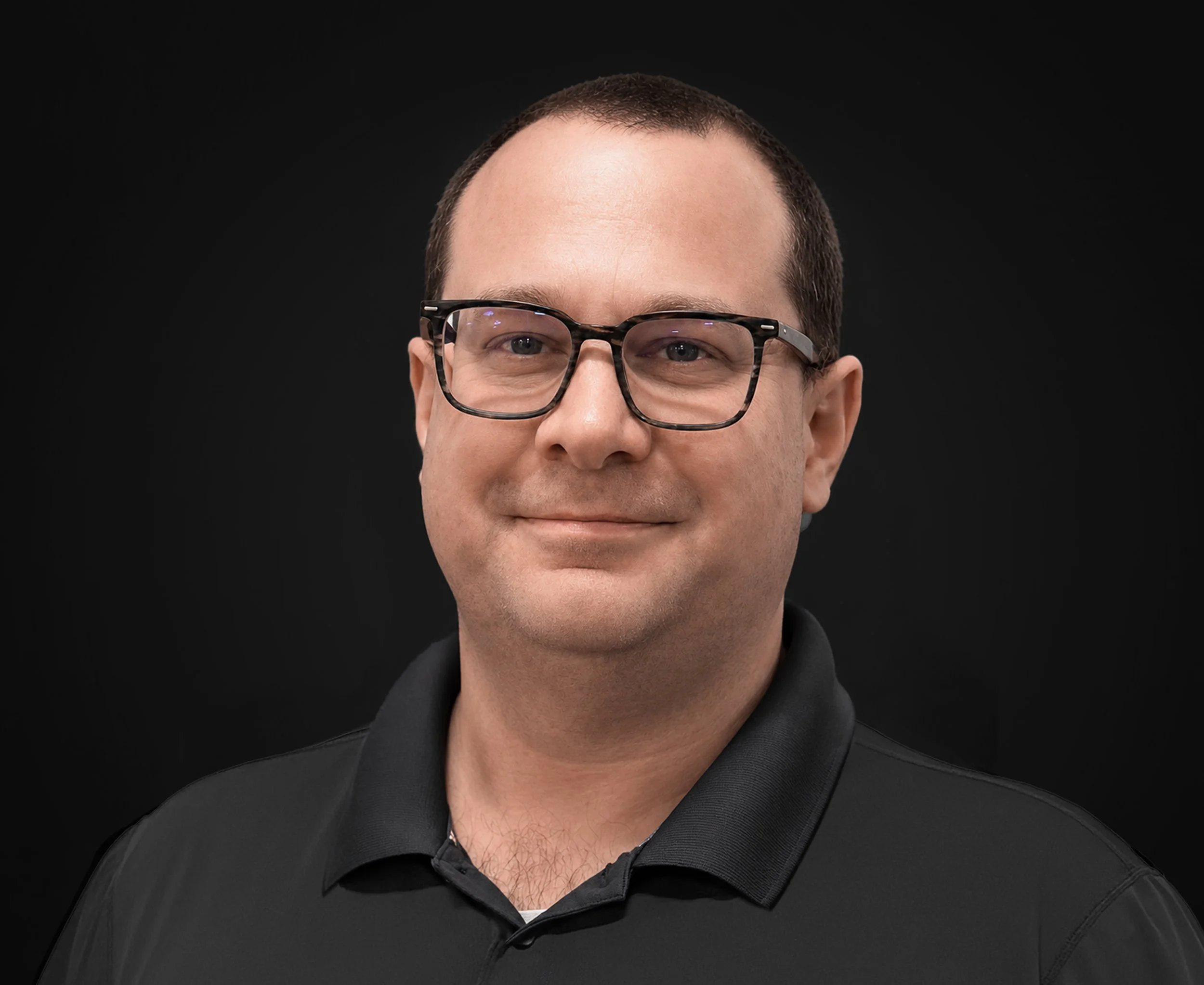 Close-up of a man with short brown hair, wearing black glasses and a black shirt, against a black background.