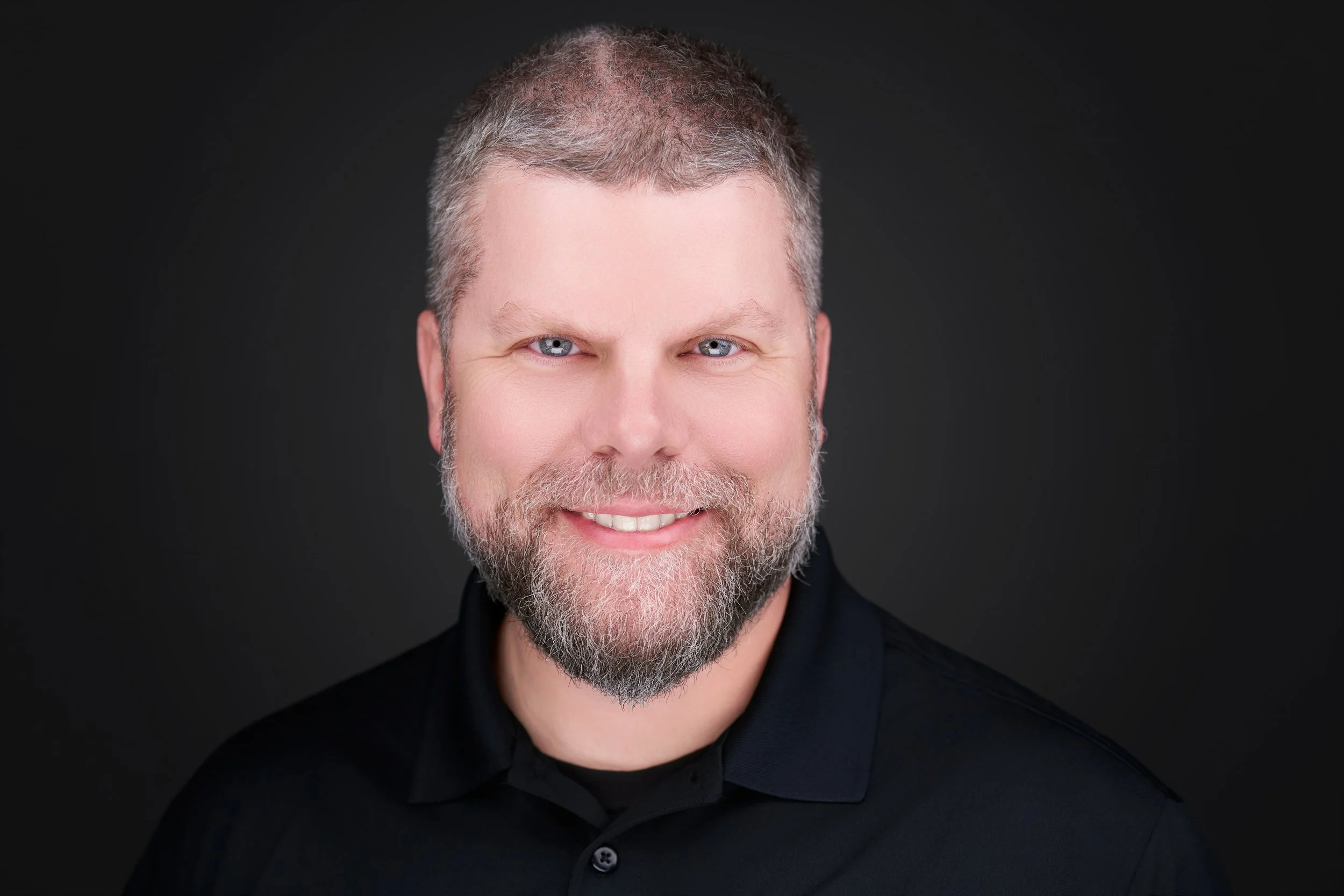 Close-up portrait of a smiling middle-aged man with gray hair and a beard, wearing a black shirt against a dark background.