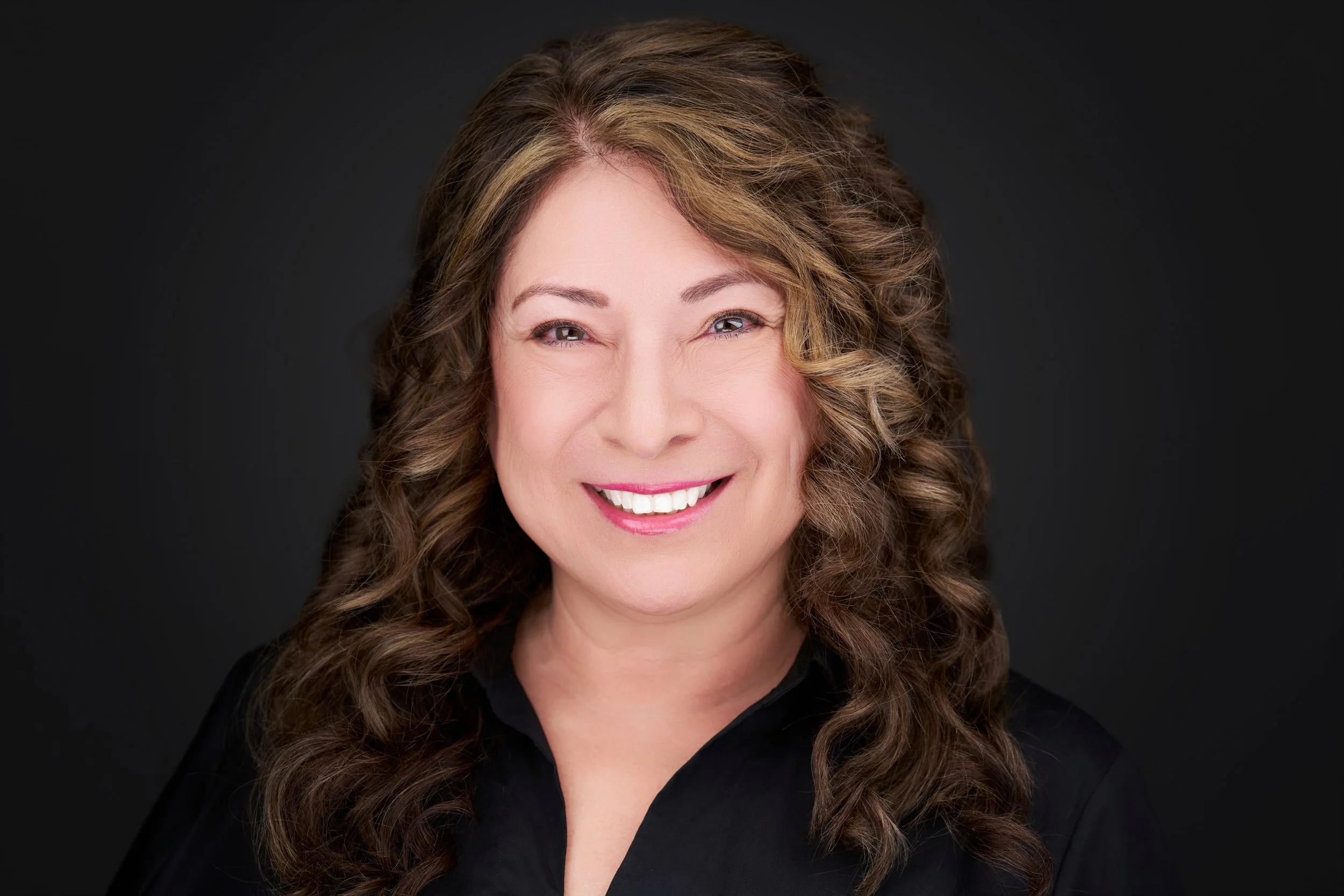 A smiling woman with wavy, light brown hair and light skin, wearing a black top, against a dark background.
