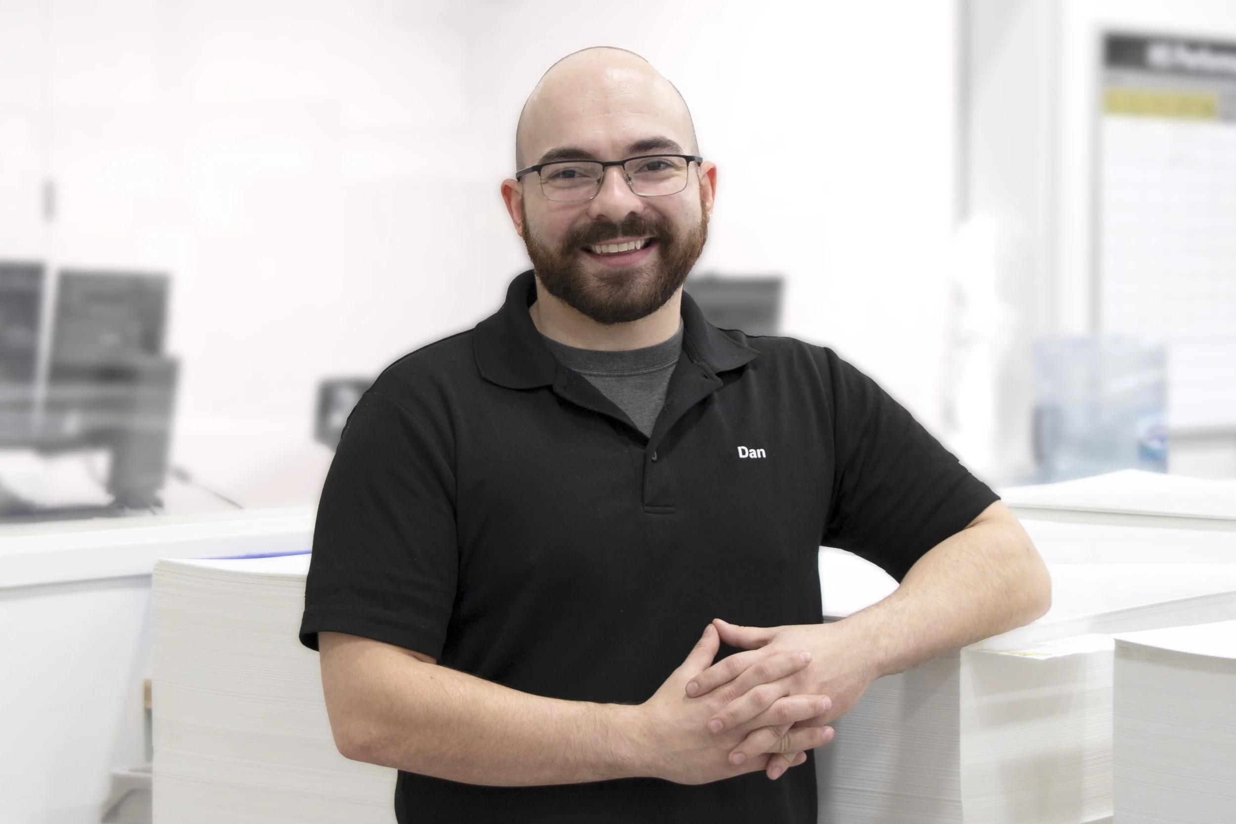 A smiling man with glasses and a beard, wearing a black polo shirt with the name "Dan" on it, sitting with arms crossed at a white table in a bright, modern office or workspace.