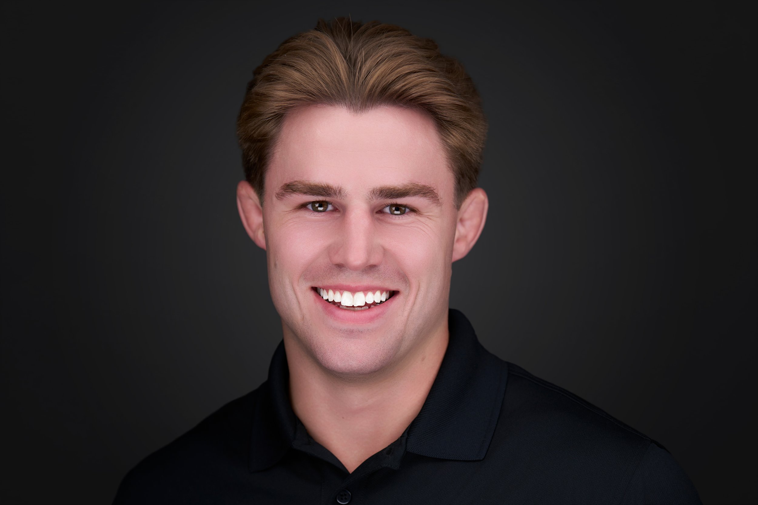 A young man with brown hair, smiling, wearing a black collared shirt, on a dark background.