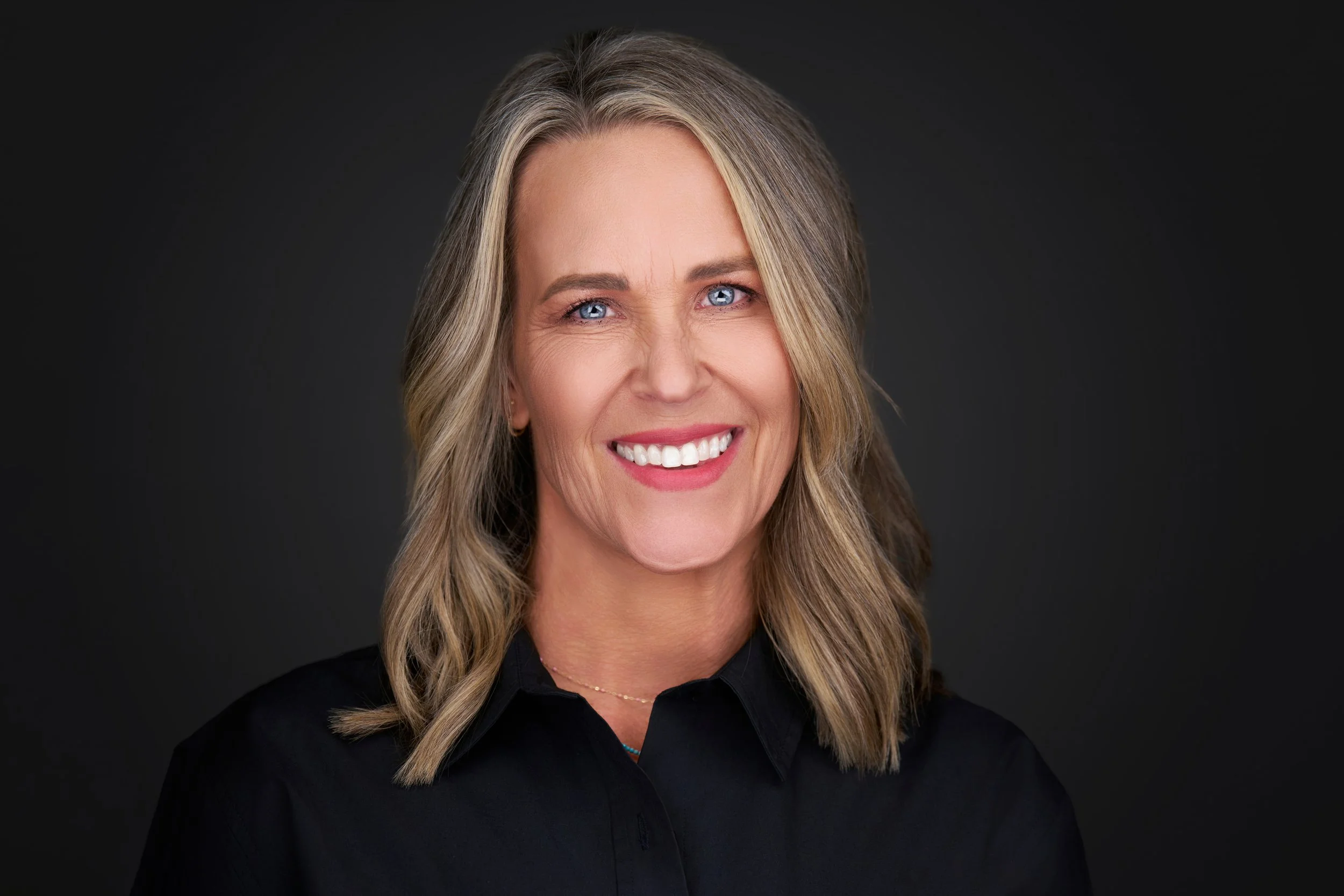 Portrait of a middle-aged woman with blonde hair, blue eyes, and a smile, wearing a black shirt against a dark background.