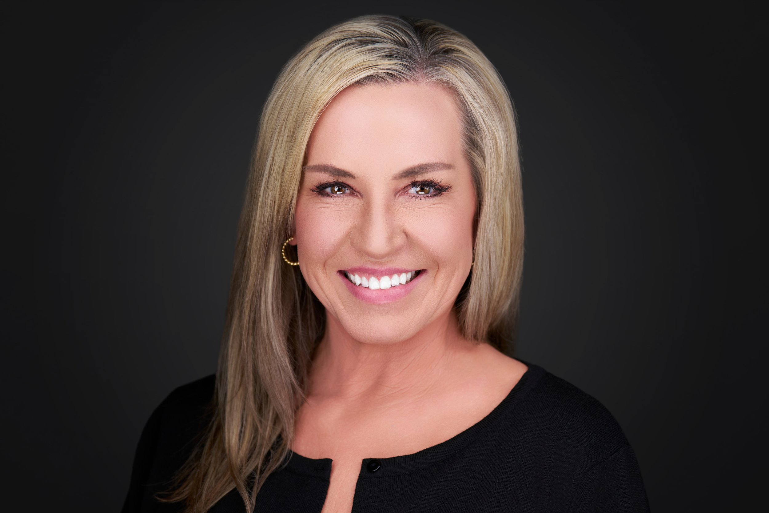 Smiling woman with blonde hair, wearing gold hoop earrings and a black top, against a dark background.