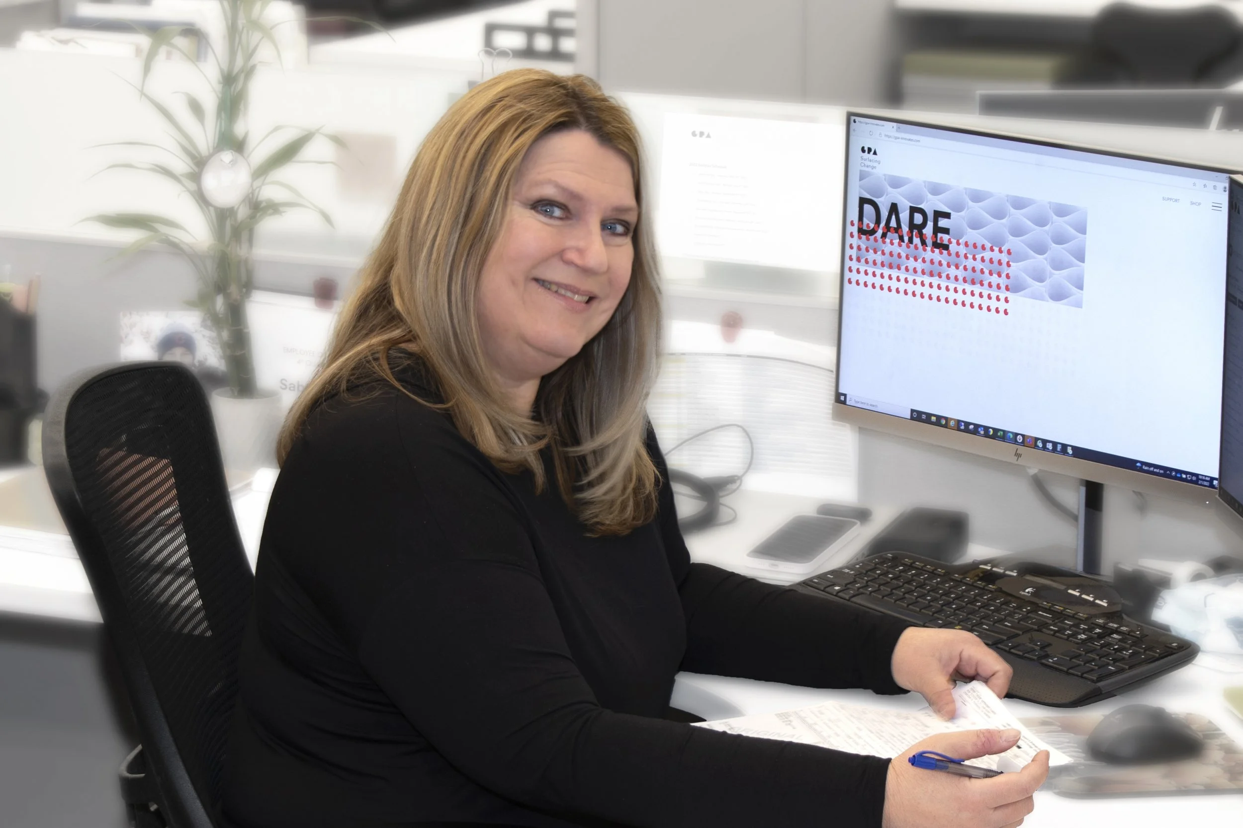 A woman with long blonde hair sitting at a desk, smiling, with a computer monitor displaying the word "DARE" and a pattern of red dots.
