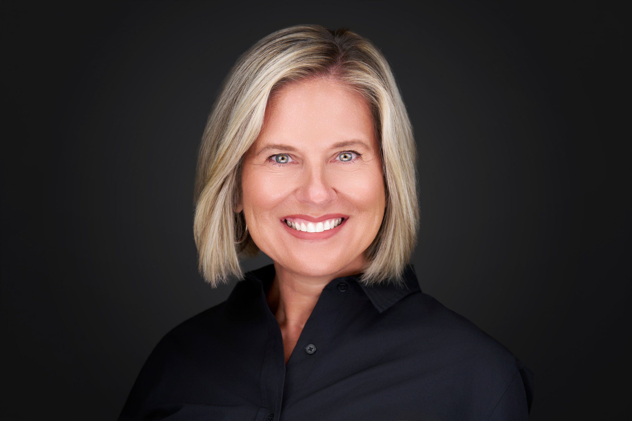 A woman with shoulder-length blonde hair, light skin, and blue eyes smiling, wearing a black shirt against a dark background. Kim Hajec
