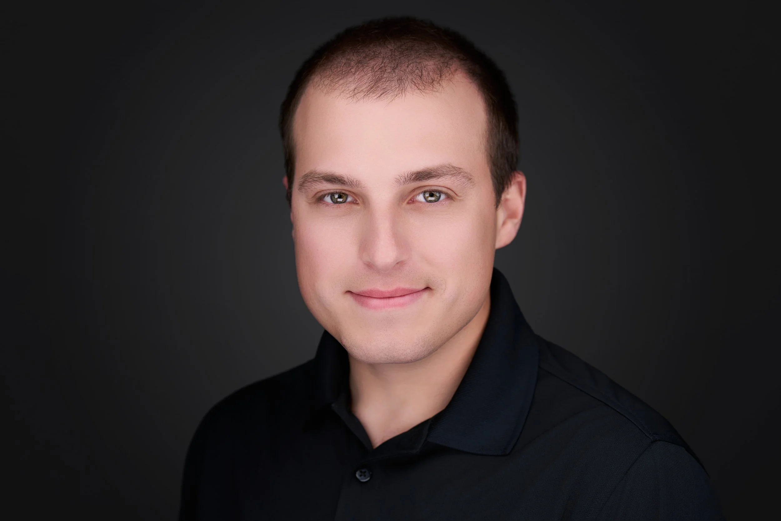 Portrait of a young man with short brown hair, hazel eyes, and a slight smile, wearing a black shirt against a dark background.