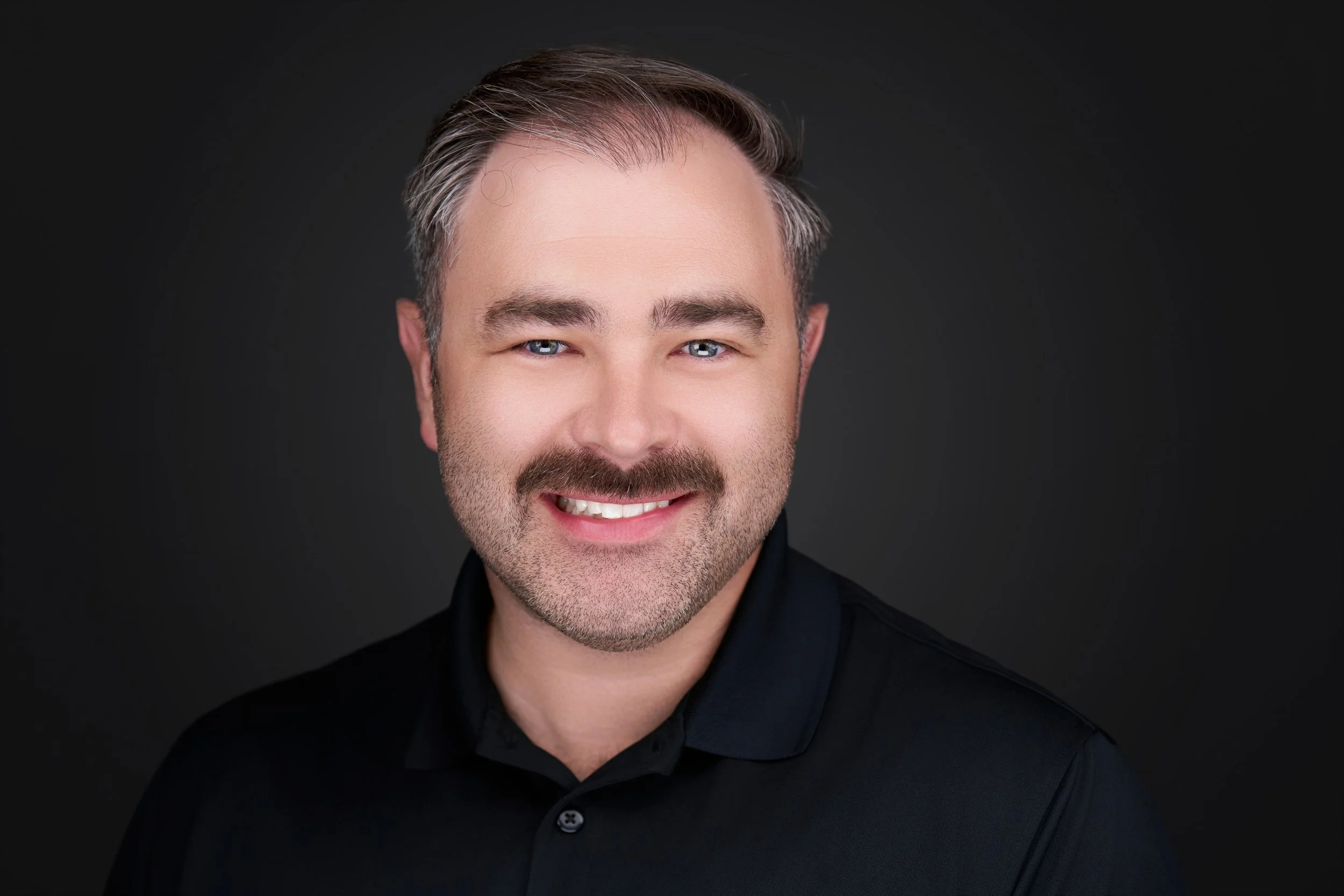 Close-up of a smiling man with dark hair, blue eyes, and a beard, wearing a black shirt against a dark background.