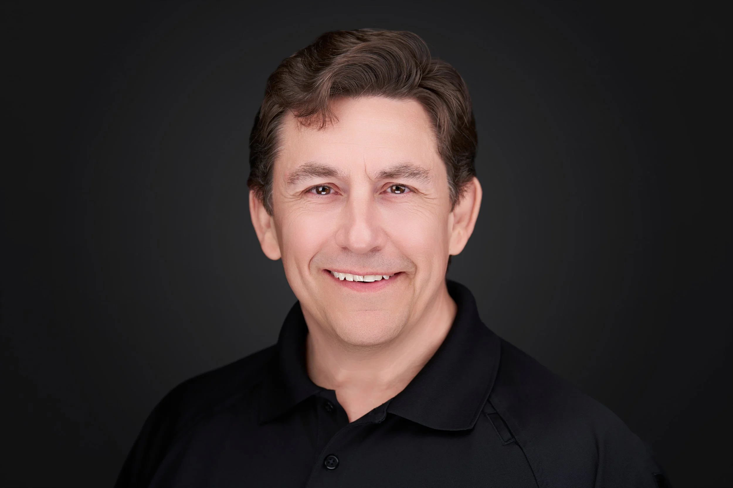 Close-up portrait of a smiling middle-aged man with brown hair and hazel eyes, wearing a black collared shirt against a dark background. Ron Gasior