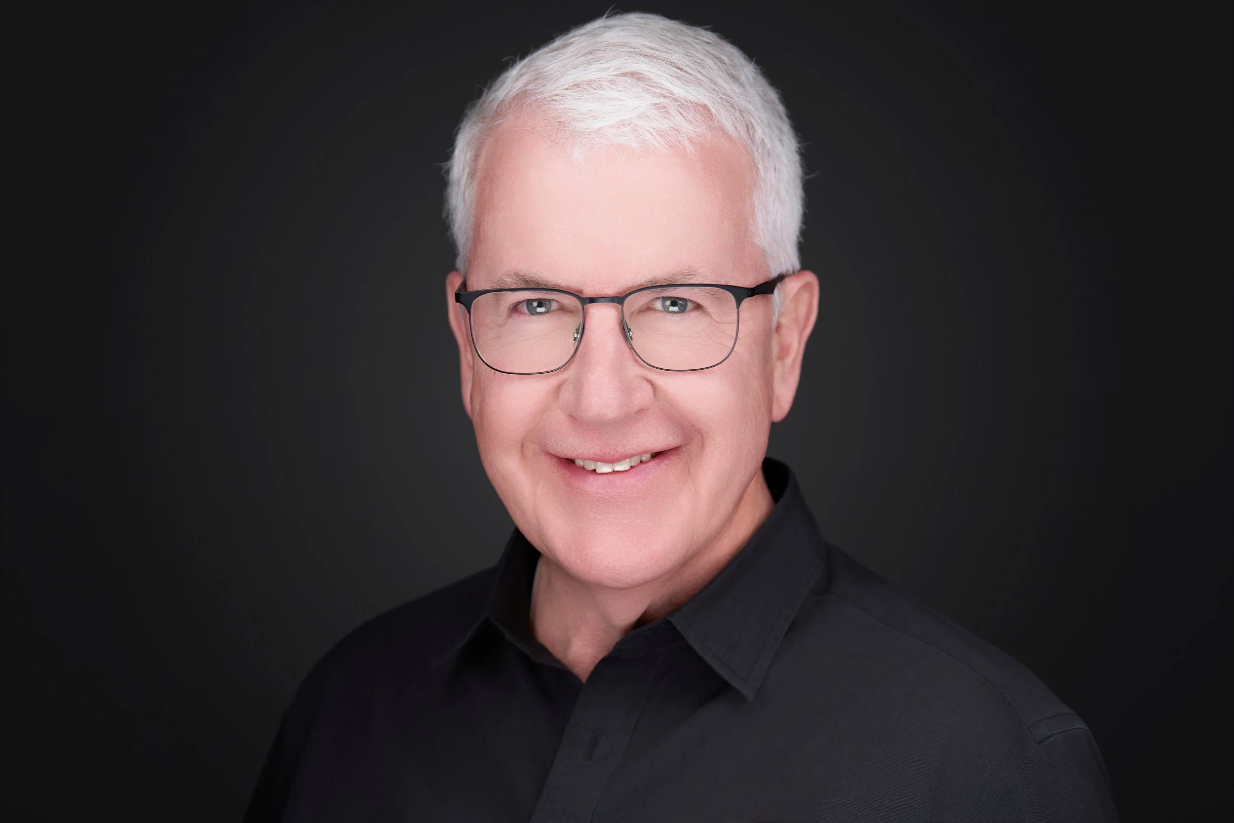 Portrait of an older man with short white hair, glasses, and a friendly expression, wearing a black shirt against a dark background.